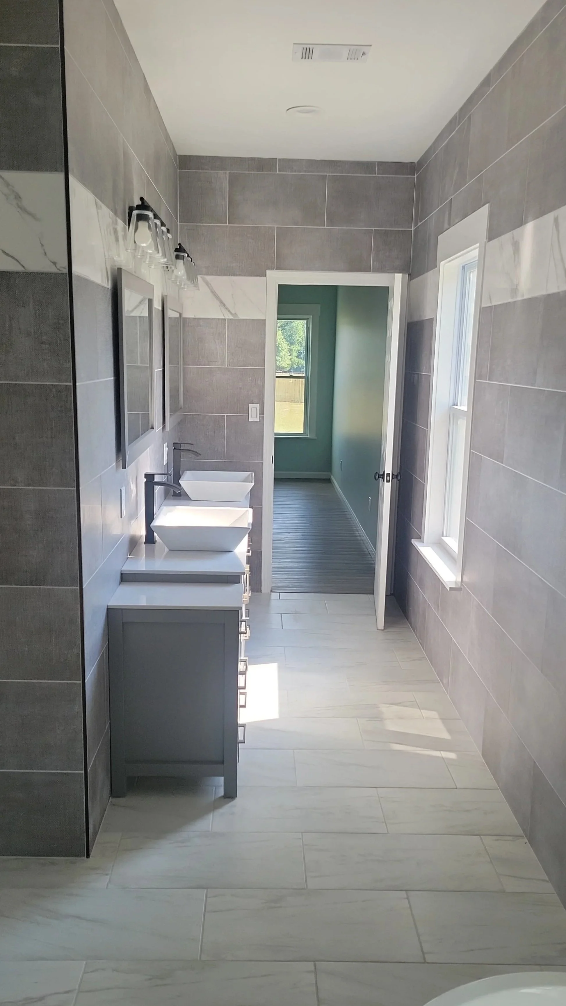 Modern bathroom with gray tiled walls, double white vessel sinks, black faucets, mirror above sinks, wall-mounted light fixtures, white floor tiles, and an open door leading to a room with green walls and wooden flooring.