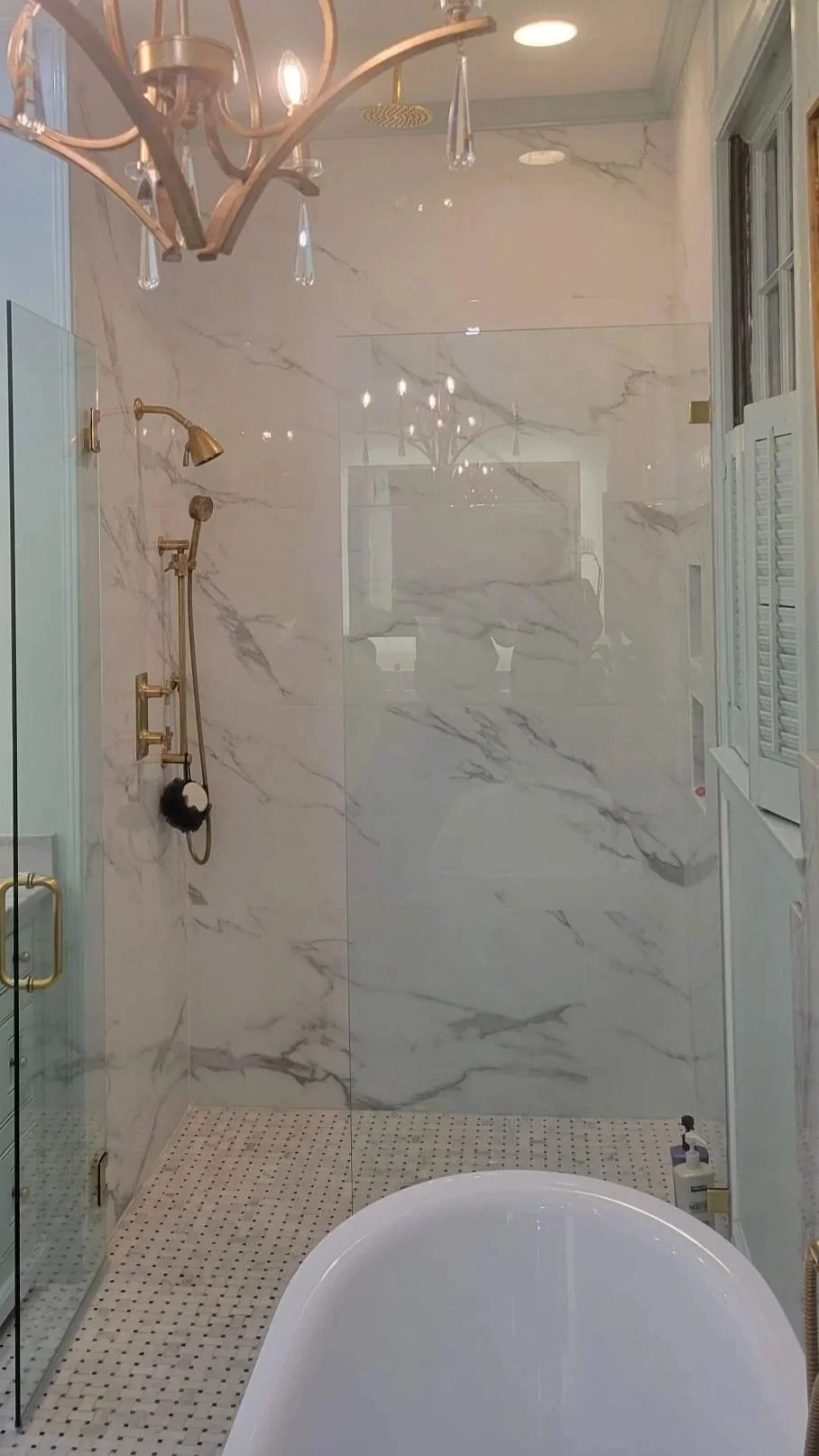 A luxurious bathroom with a marble shower, gold fixtures, a chandelier, and a bathtub in the foreground.