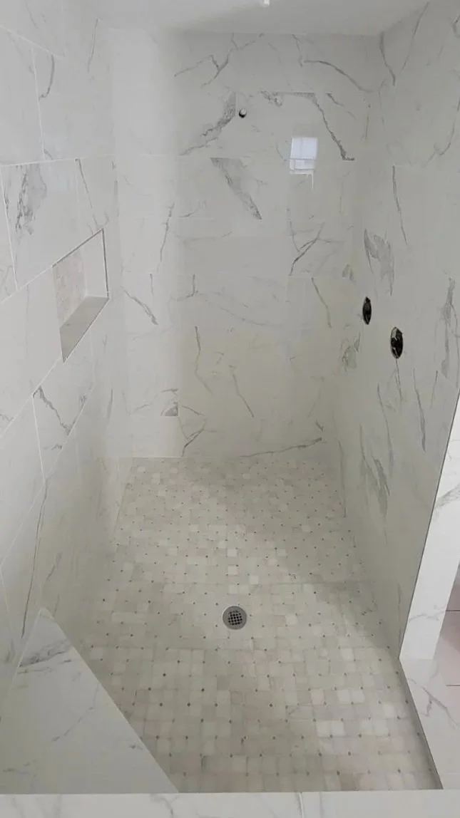Empty shower stall with marble-like tiled walls and a small drain at the floor, with an unfinished ceiling fixture.