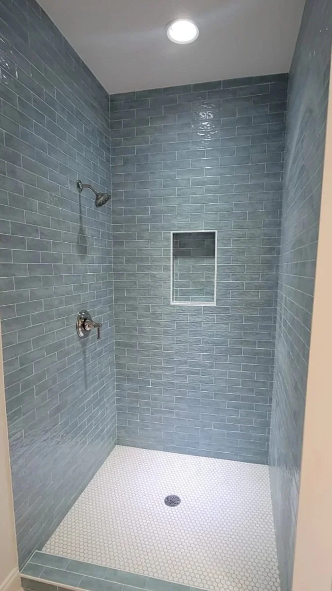 A walk-in shower with gray subway tiles on the walls, a built-in niche, a ceiling light, and a white hexagon tile floor, with an exterior tiled threshold.