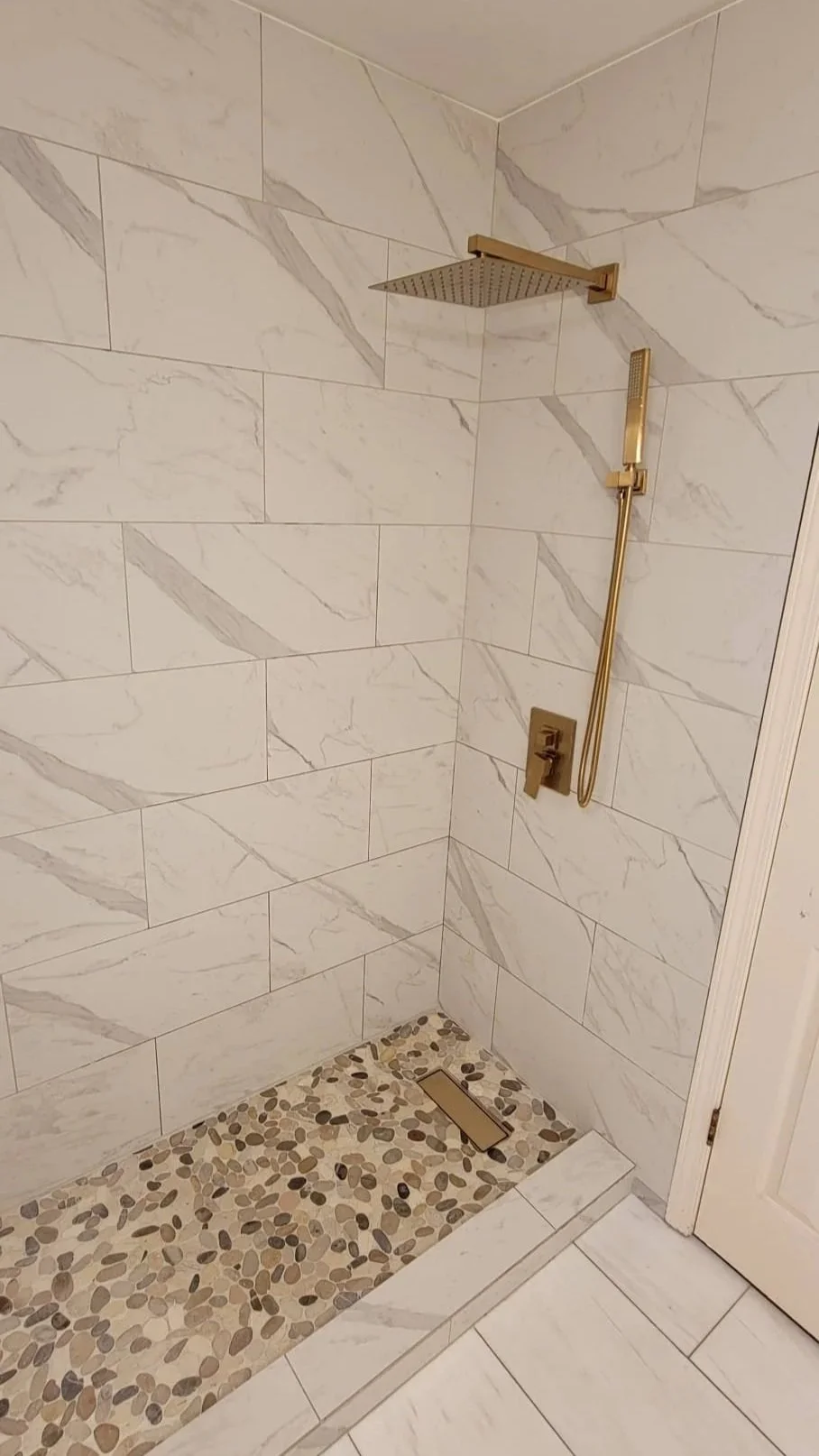 Shower with white marble tiles, gold fixtures, and a pebble stone shower floor with a drain.