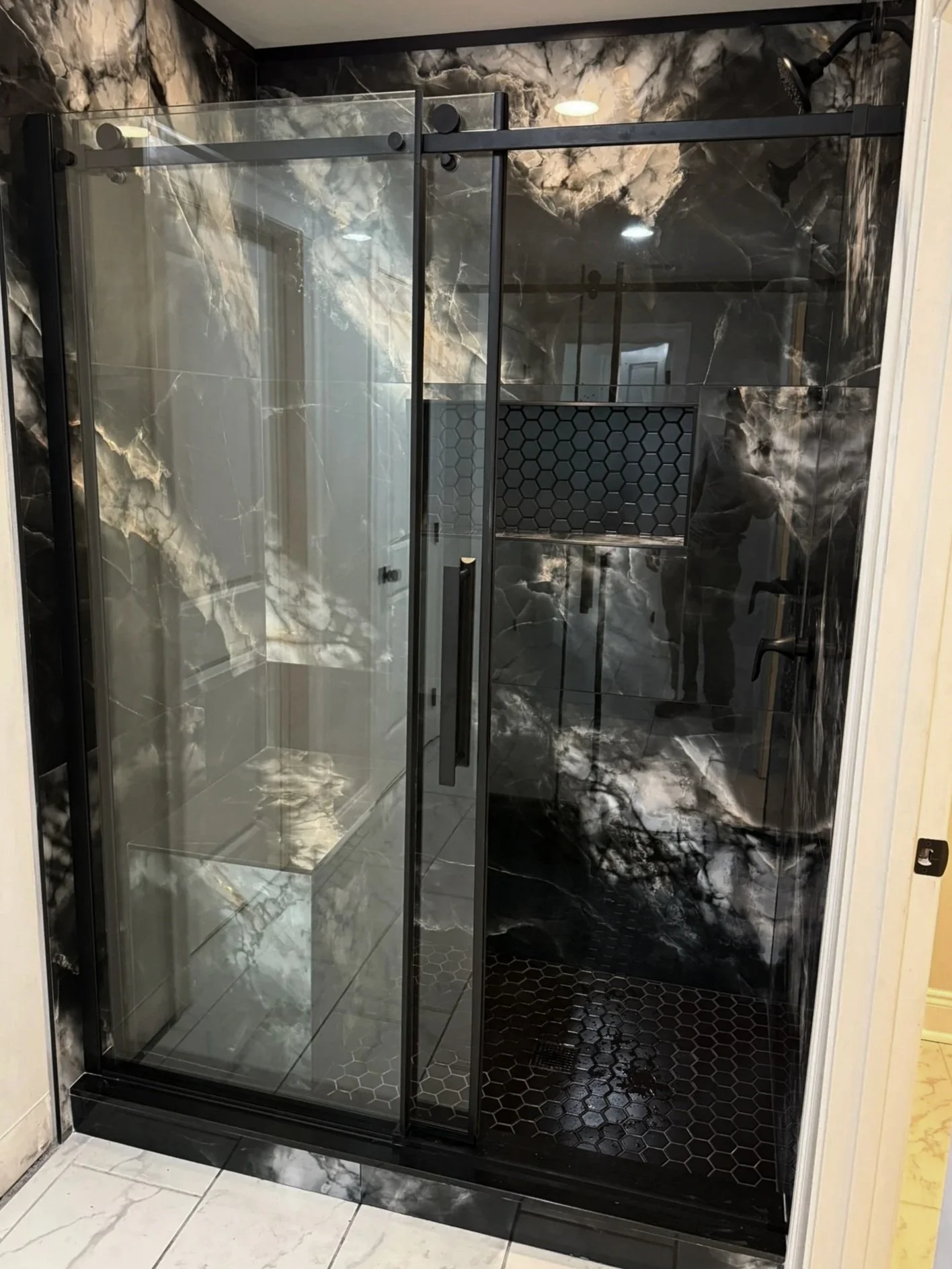 A modern shower enclosure with black metal framing, a glass door, and marble-look wall tiles in black, white, and gray tones, with black hexagonal floor tiles and a built-in niche.