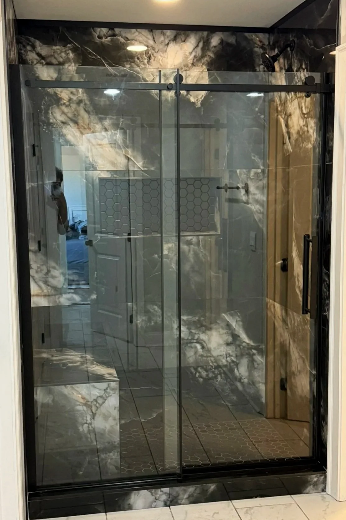 A modern walk-in shower with dark gray and white marble tiles, a glass sliding door, black hardware, and a ceiling light