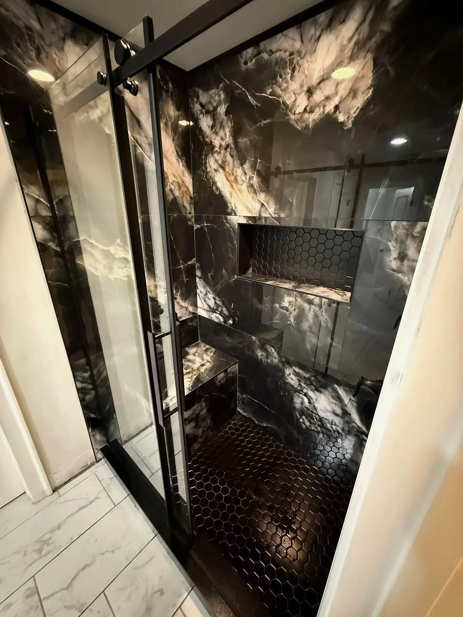 A modern bathroom shower area with black marble walls featuring white and gold veining, a built-in niche with honeycomb tile, a black hexagon tiled floor, and a glass door with black metal hardware.