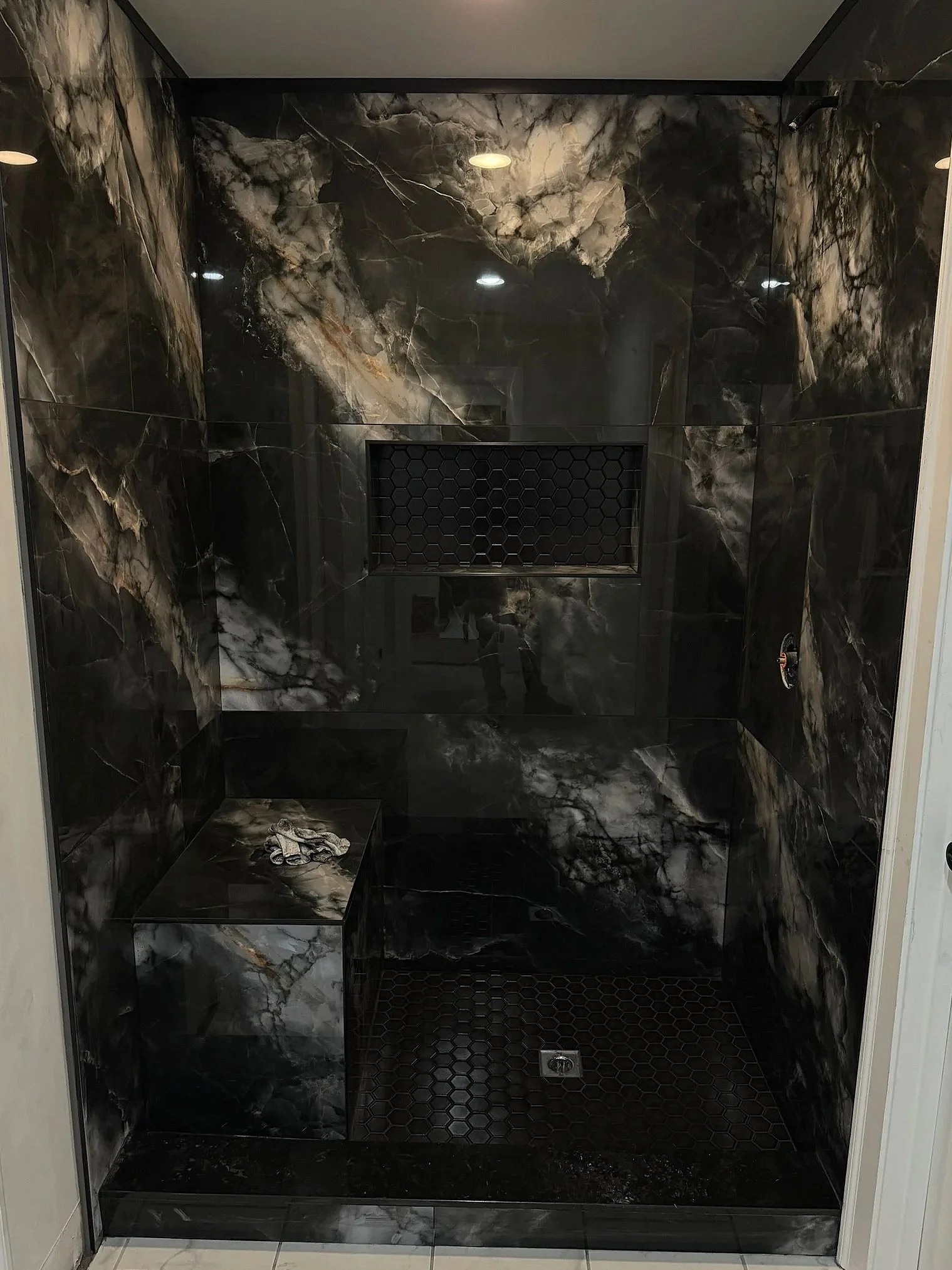 A luxurious shower stall with black marble-like tiles with white and gray veining, a built-in bench, and a black honeycomb tile vent on the back wall.