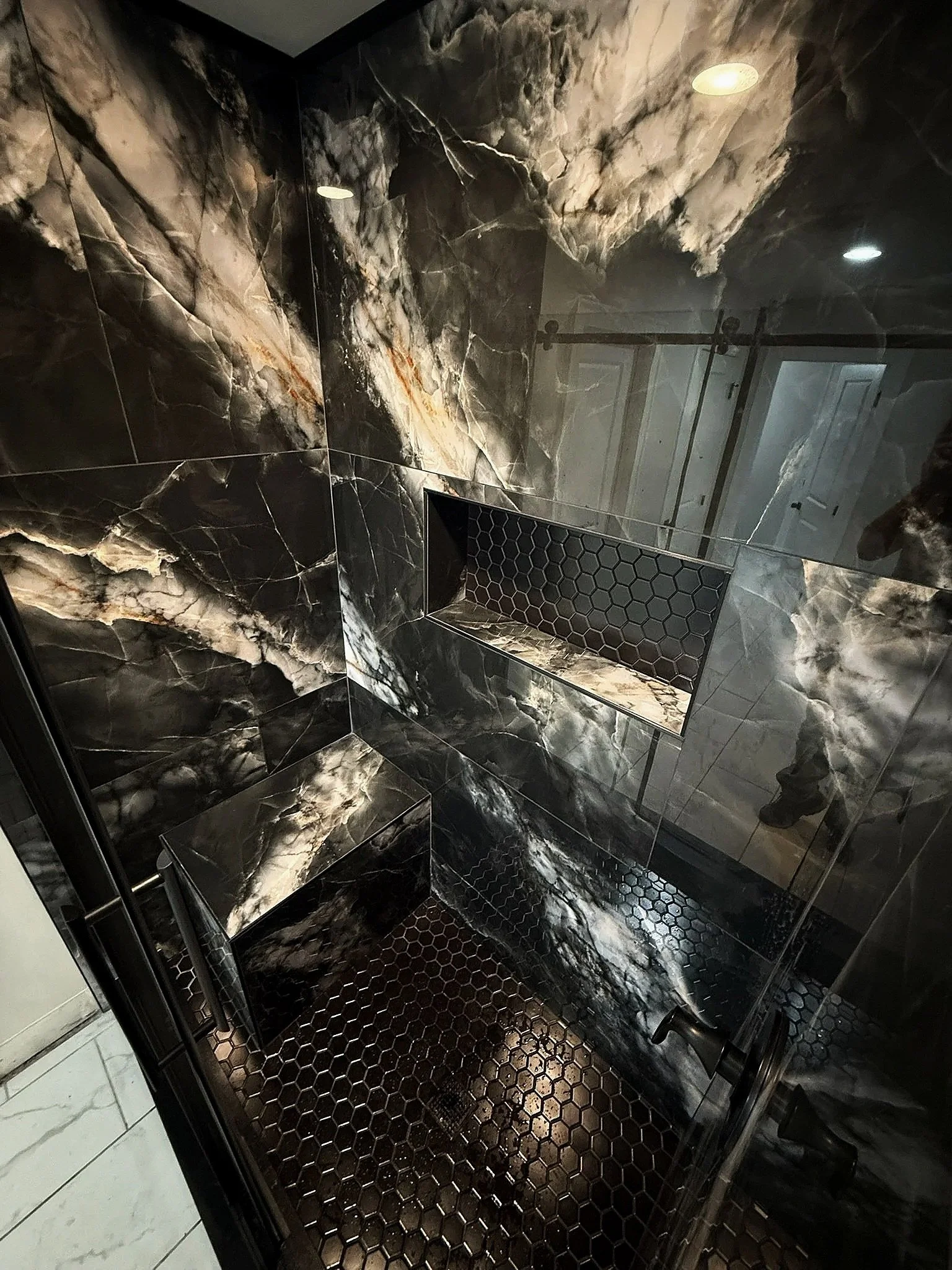 A shower stall with black and white marble walls, a black hexagonal tiled niche, and a black hexagonal tiled floor.