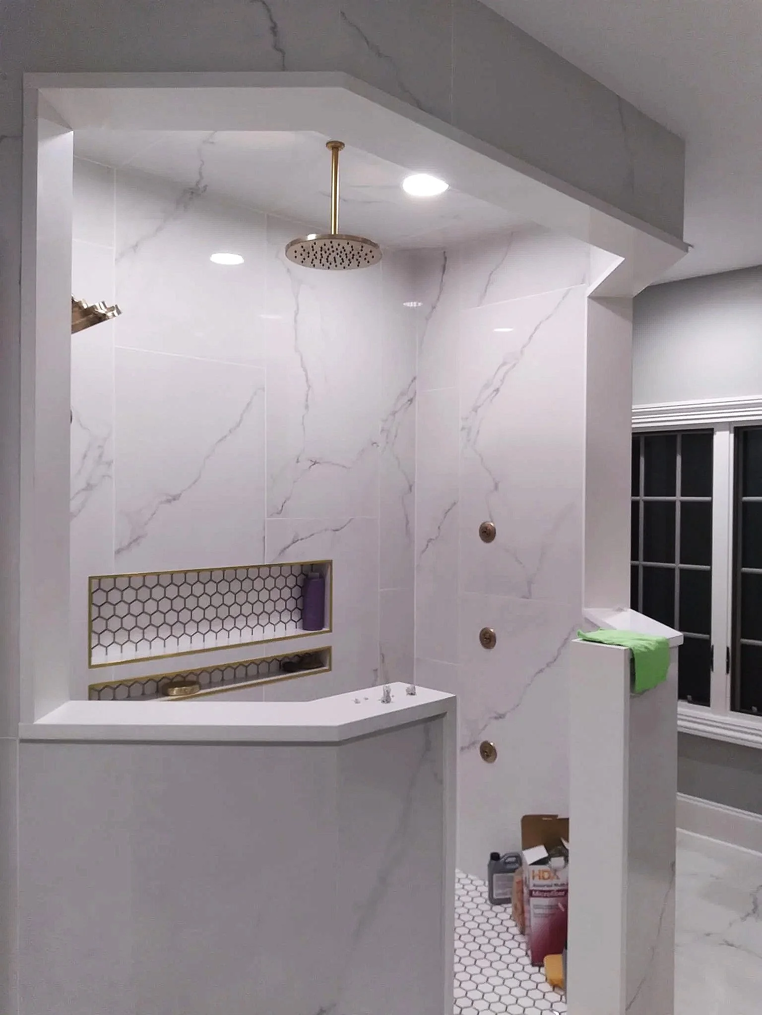 A modern bathroom with a marble shower wall, ceiling-mounted rainfall showerhead, built-in niches, and a small partition with a green towel next to a window.