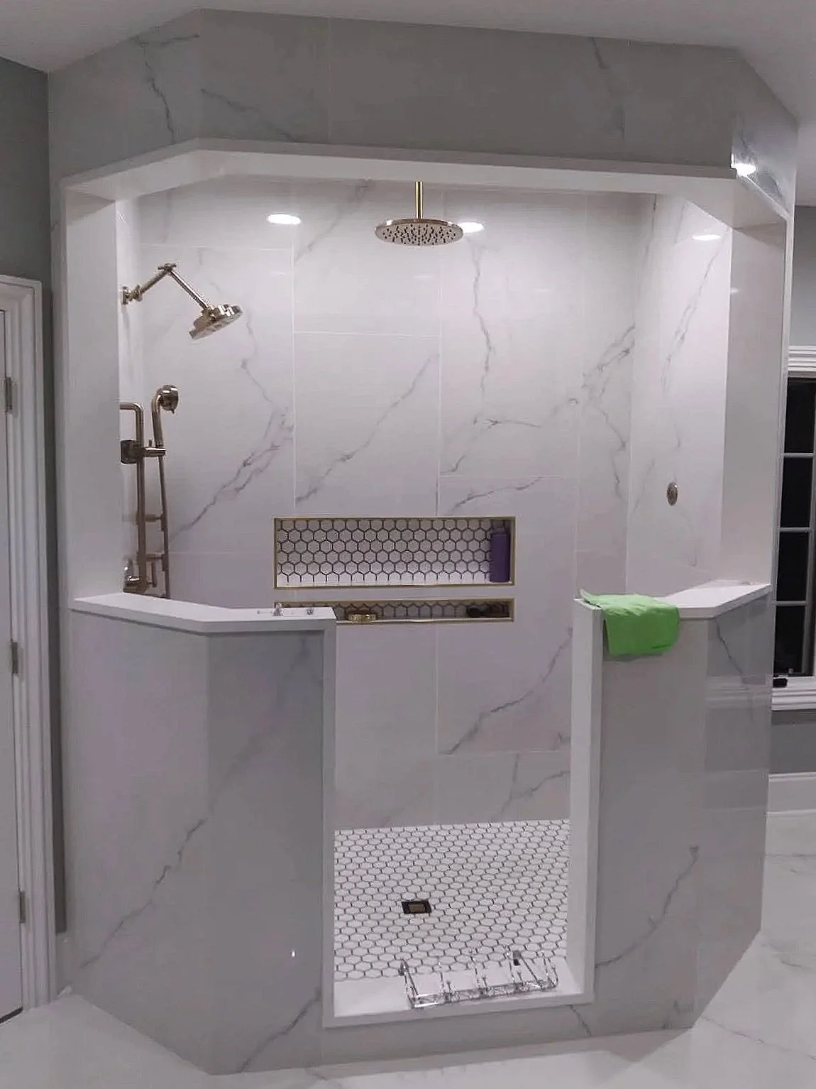 Modern shower with white marble tiles, a rain showerhead, a second wall-mounted showerhead, a built-in niche with hexagonal tiles, and a partial glass door.