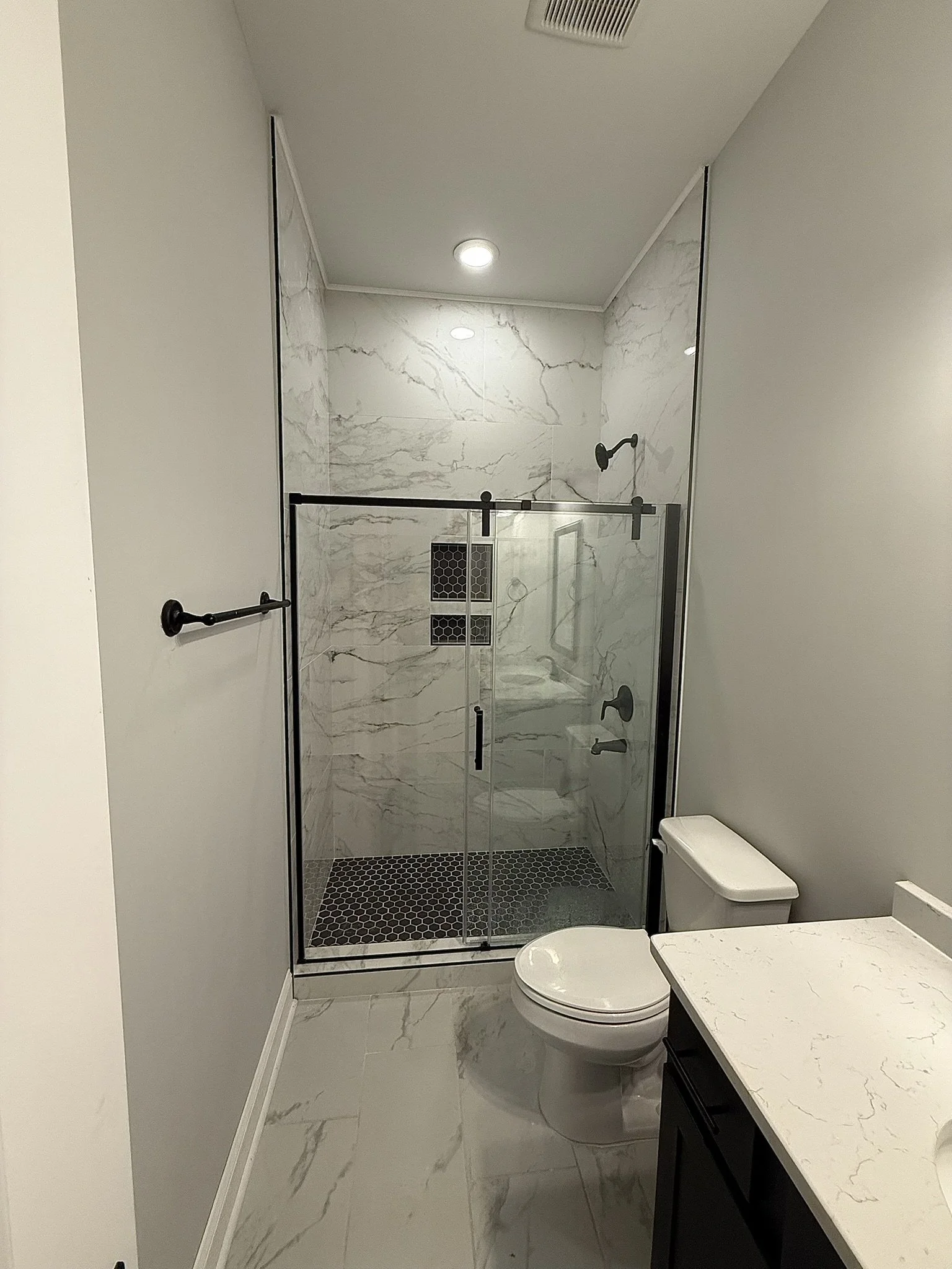A modern bathroom with a marble shower enclosure, a white toilet, a white marble countertop with a black cabinet, and gray walls. The shower has black hardware and small hexagonal black tiles on the floor.