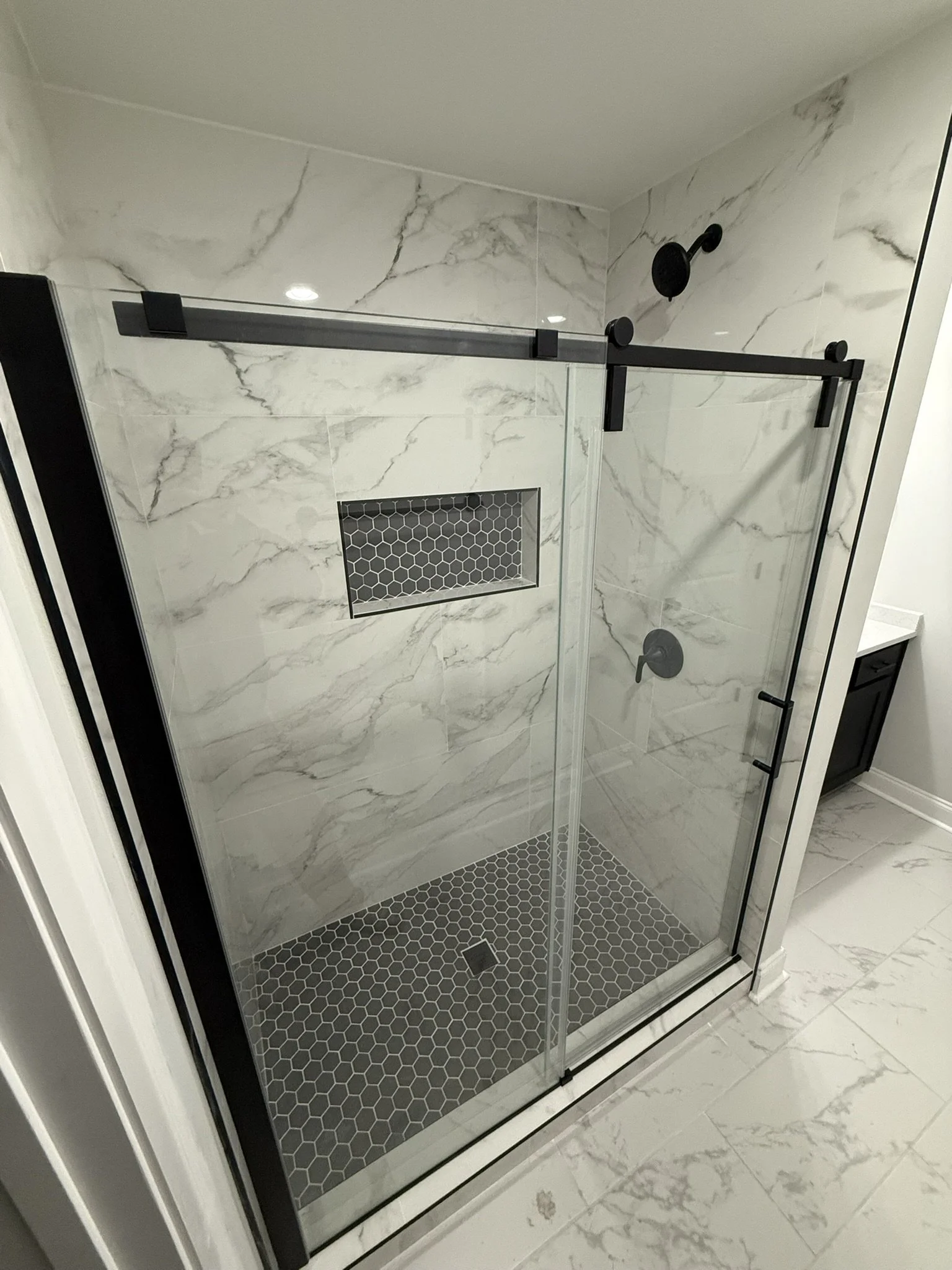 Modern walk-in shower with glass sliding door, black hardware, gray marble walls, black hexagon tile floor, recessed niche with black hex tiles, and black shower fixtures.