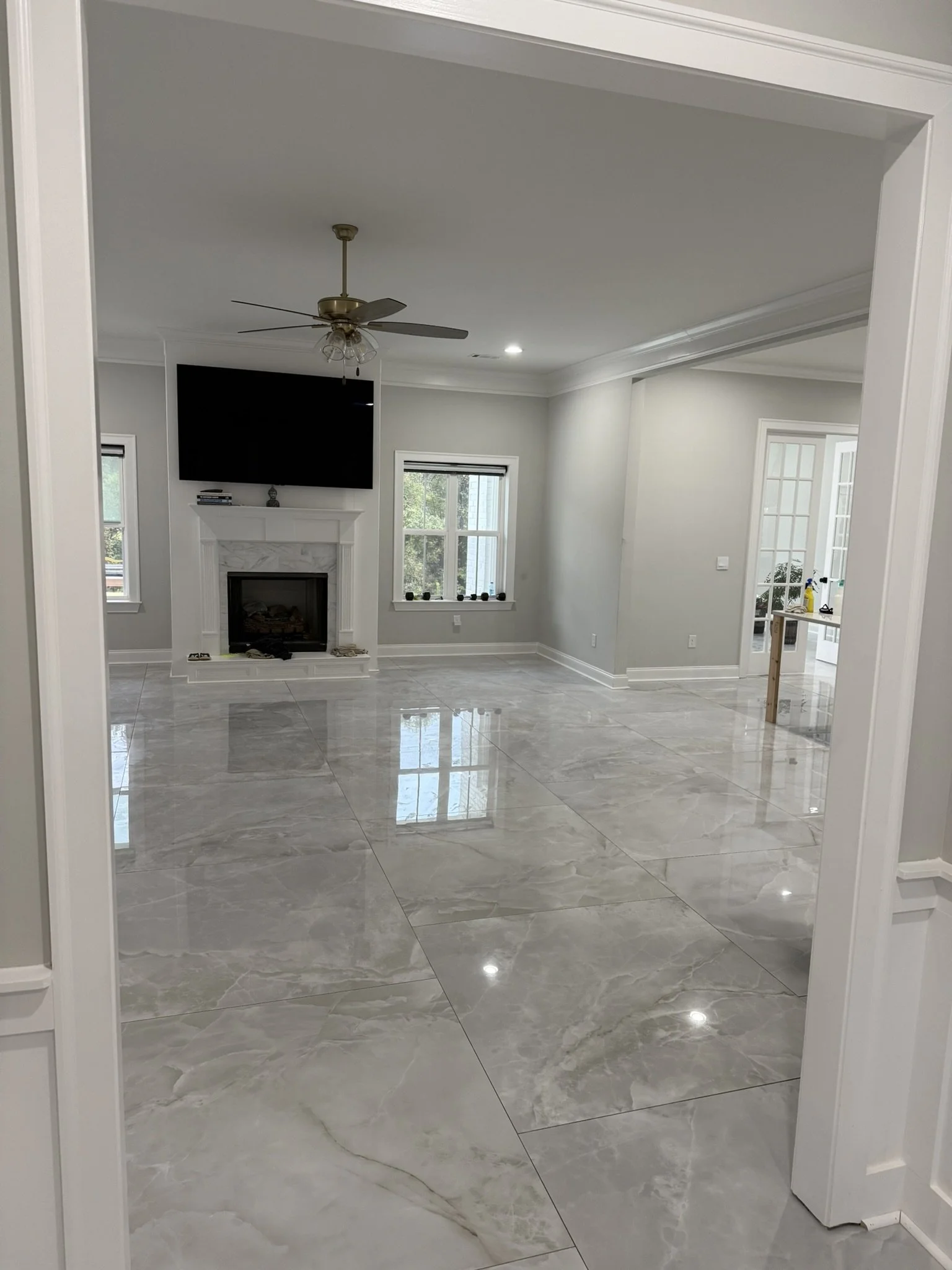 Empty living room with marble flooring, fireplace with mantel, wall-mounted TV, two windows, ceiling fan, and adjacent sunroom with glass door.