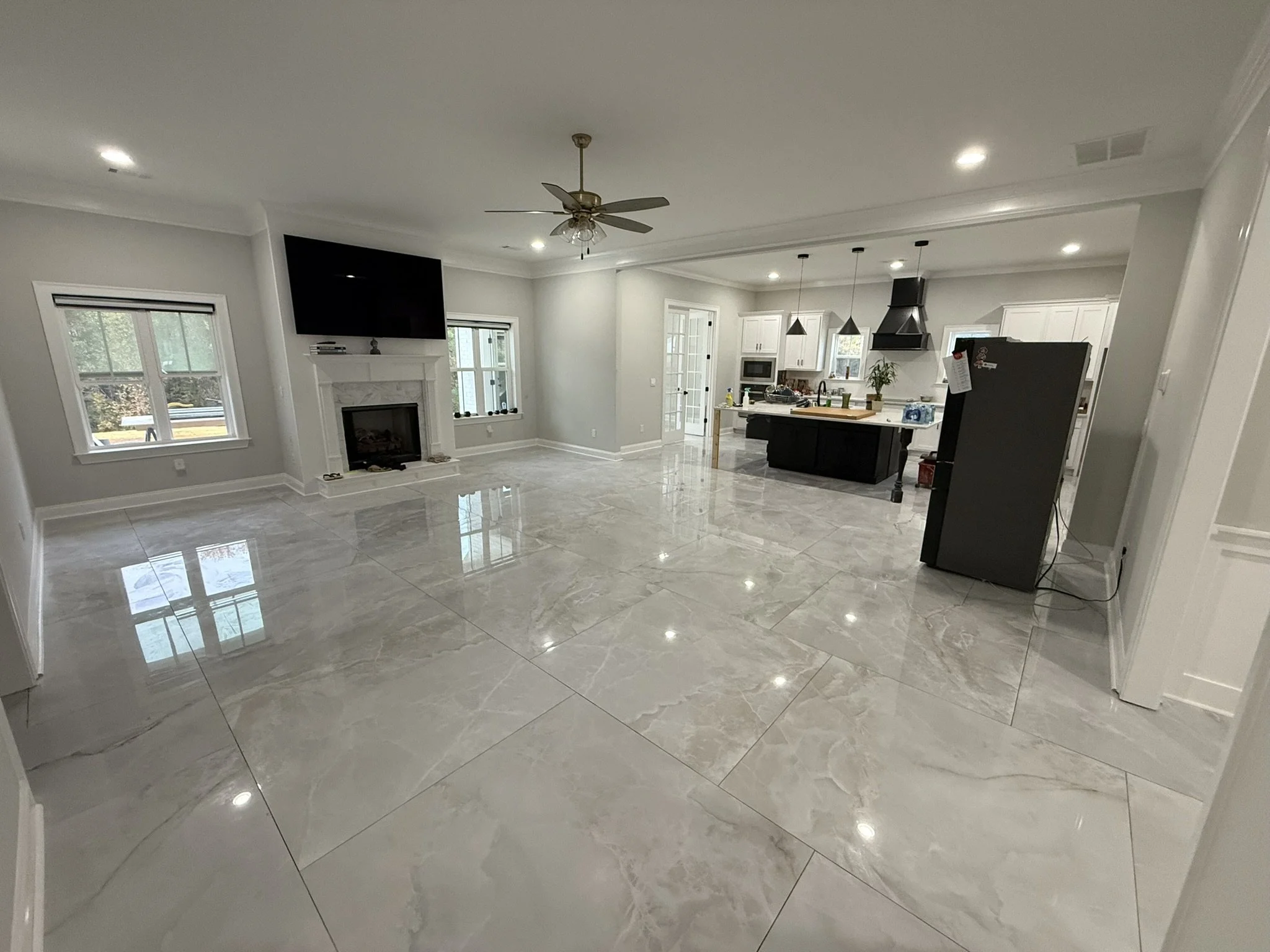 Open-concept living room and kitchen with tile flooring, white walls, windows, a ceiling fan, a fireplace, a large flat-screen TV mounted above the fireplace, and a black refrigerator.