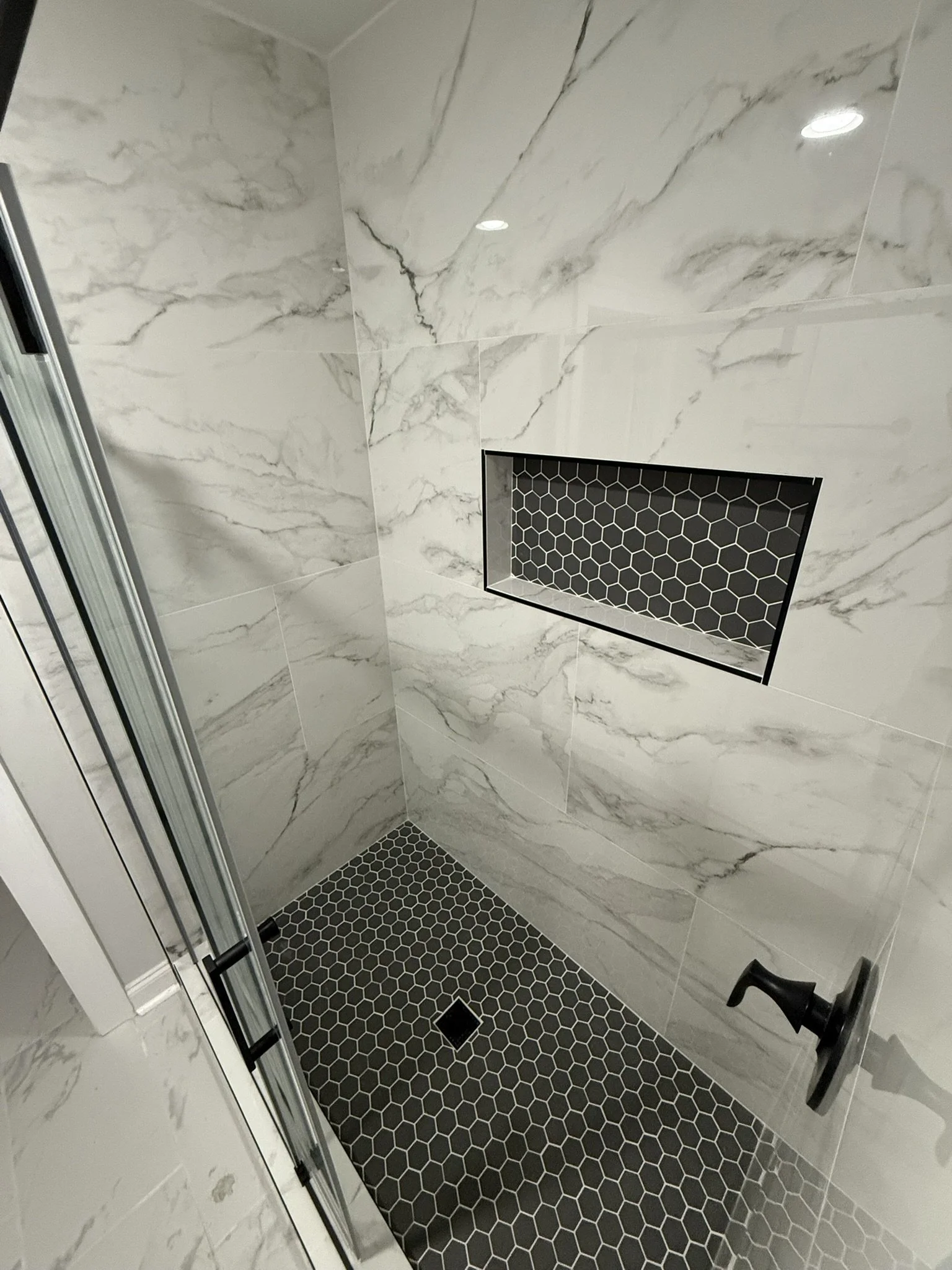 A modern shower stall with white marble walls, a black hexagonal tile floor, a recessed shelf with black honeycomb tile, and a black shower handle.
