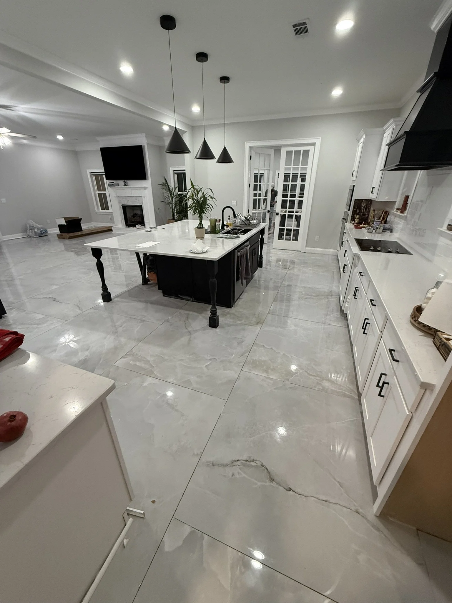 Modern kitchen with a large island countertop, white cabinetry, black hardware, marble floors, black pendant lights, and a living area with a fireplace and TV in the background.