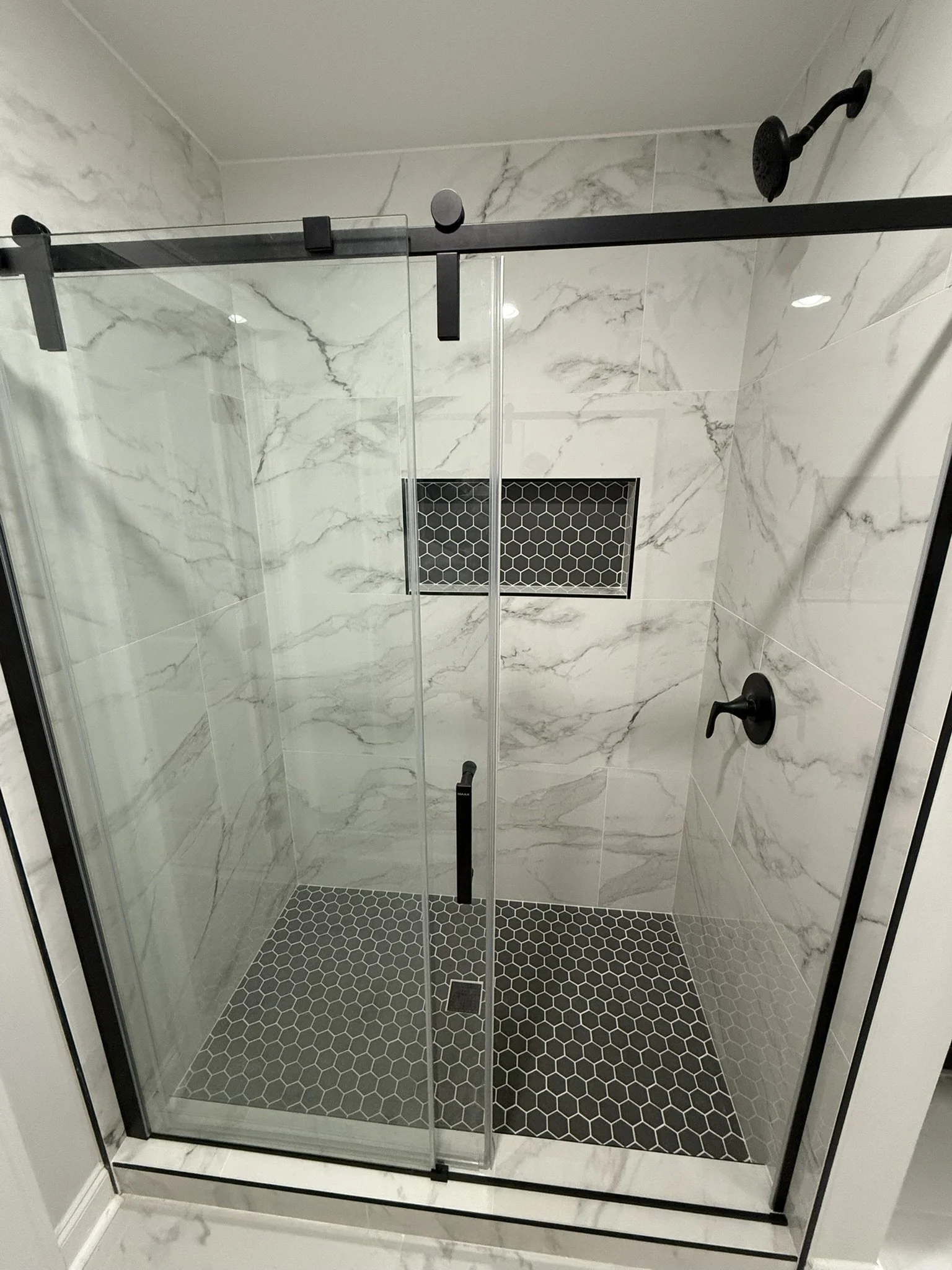 Modern walk-in shower with glass enclosure, black hardware, marble walls, black hexagon floor tiles, and built-in niche for toiletries.