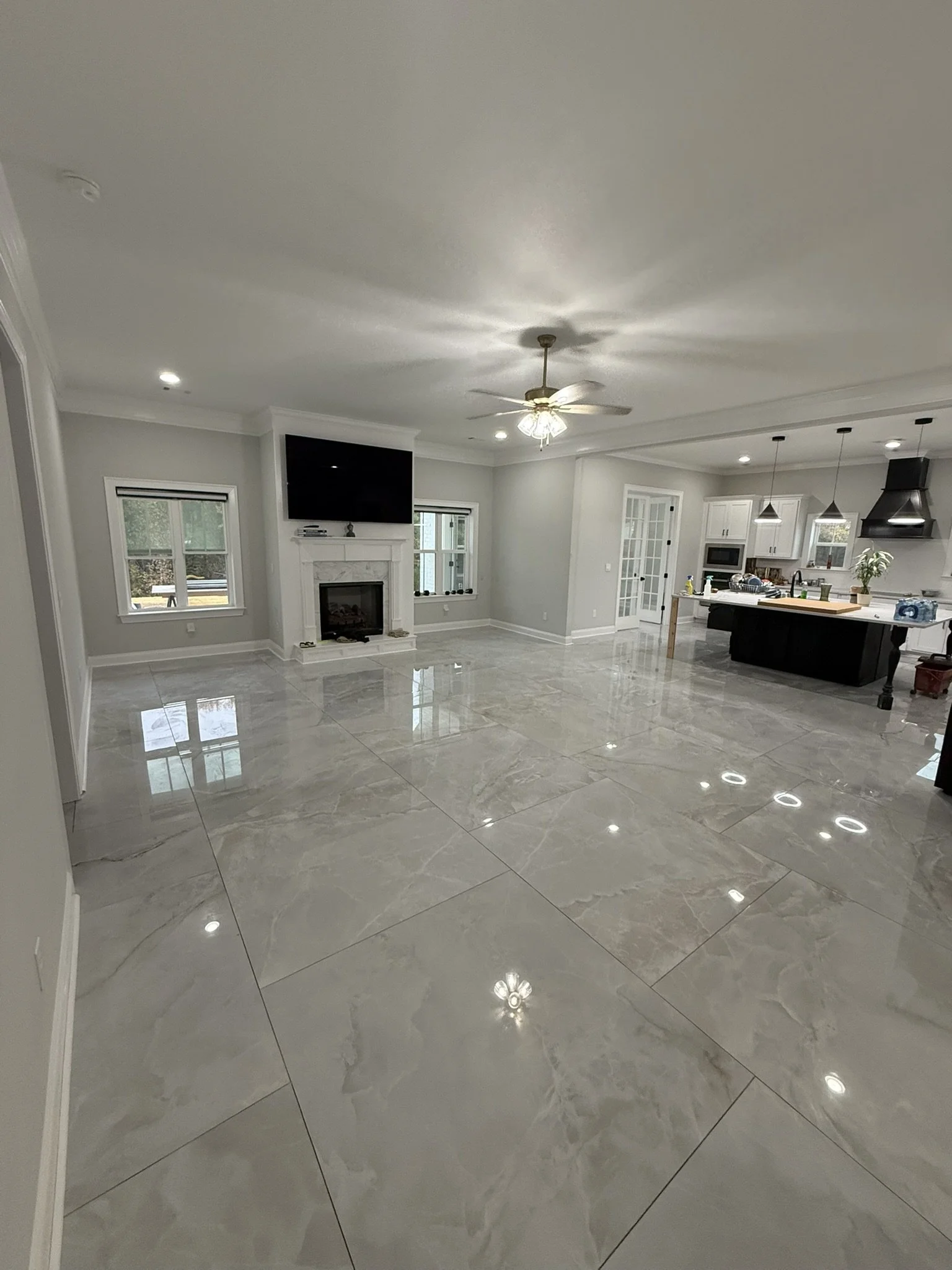 Spacious living room with glossy marble floors, a fireplace with a mounted TV above, and large windows. An open kitchen with a central island, pendant lights, and modern appliances is visible in the background.