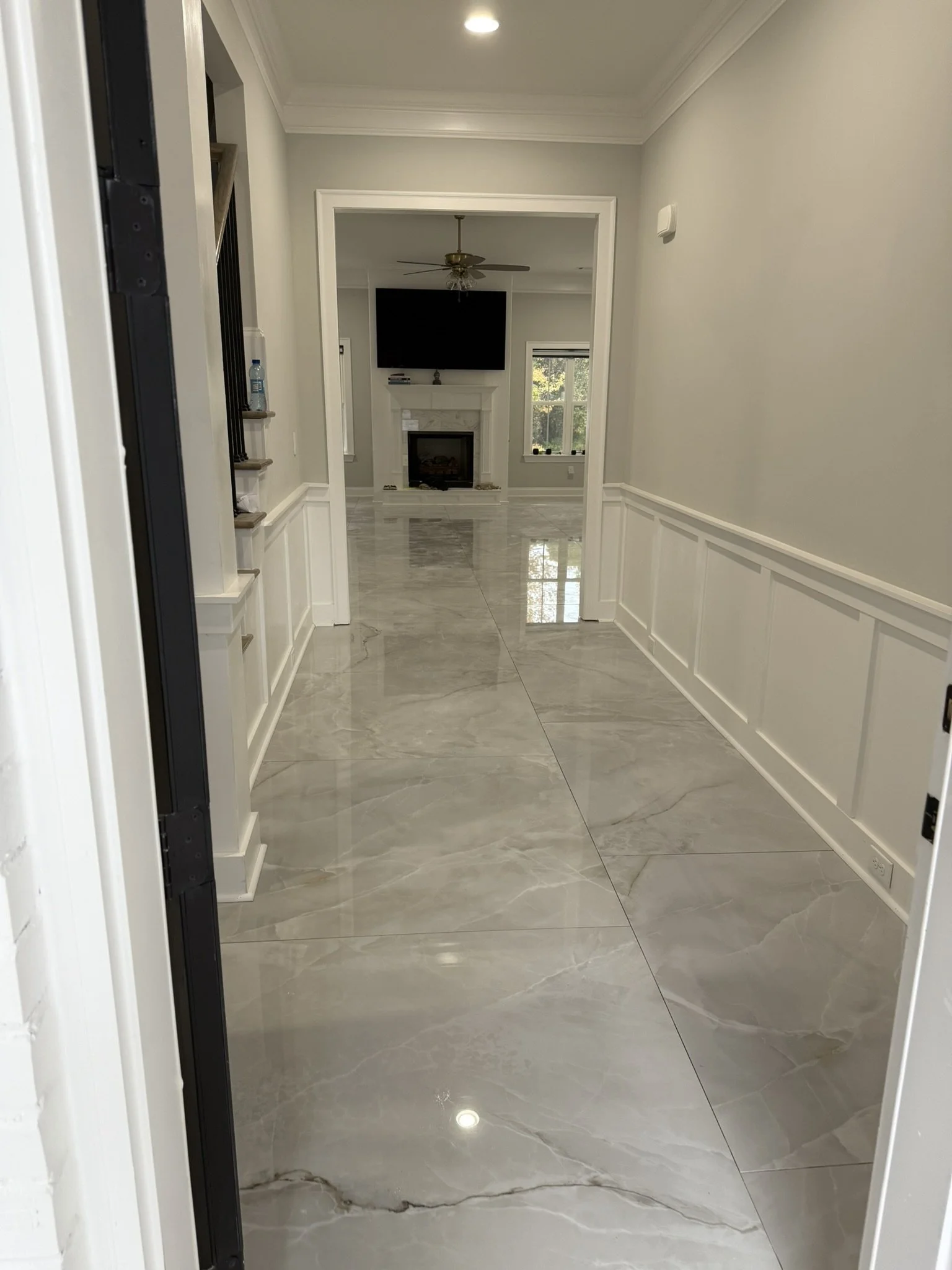 View down a hallway leading to a living room with a fireplace, TV, ceiling fan, large window, and marble flooring.