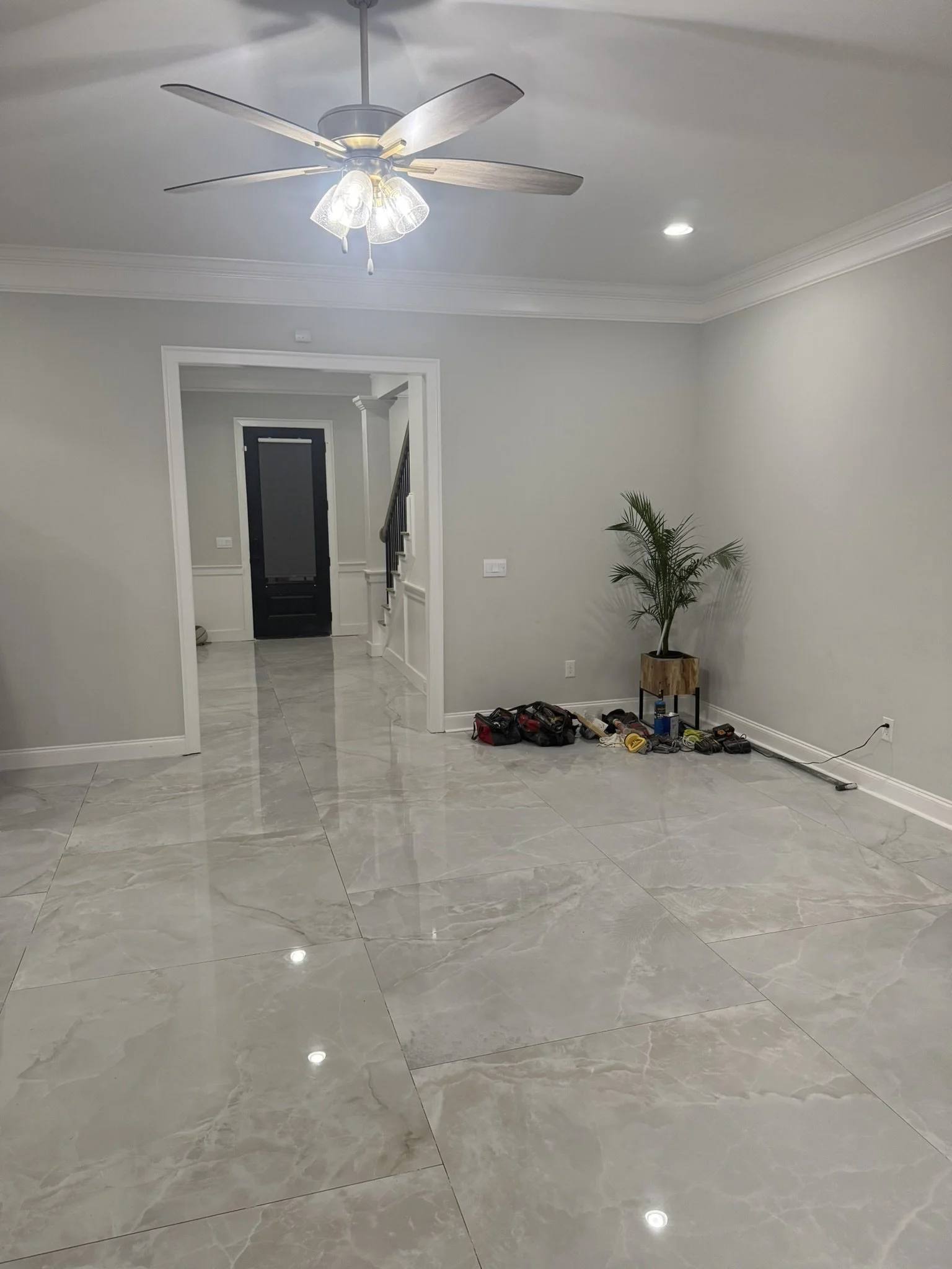 An empty living room with large marble tile flooring, a ceiling fan with lights, a potted plant, and some tools on the floor near the wall.