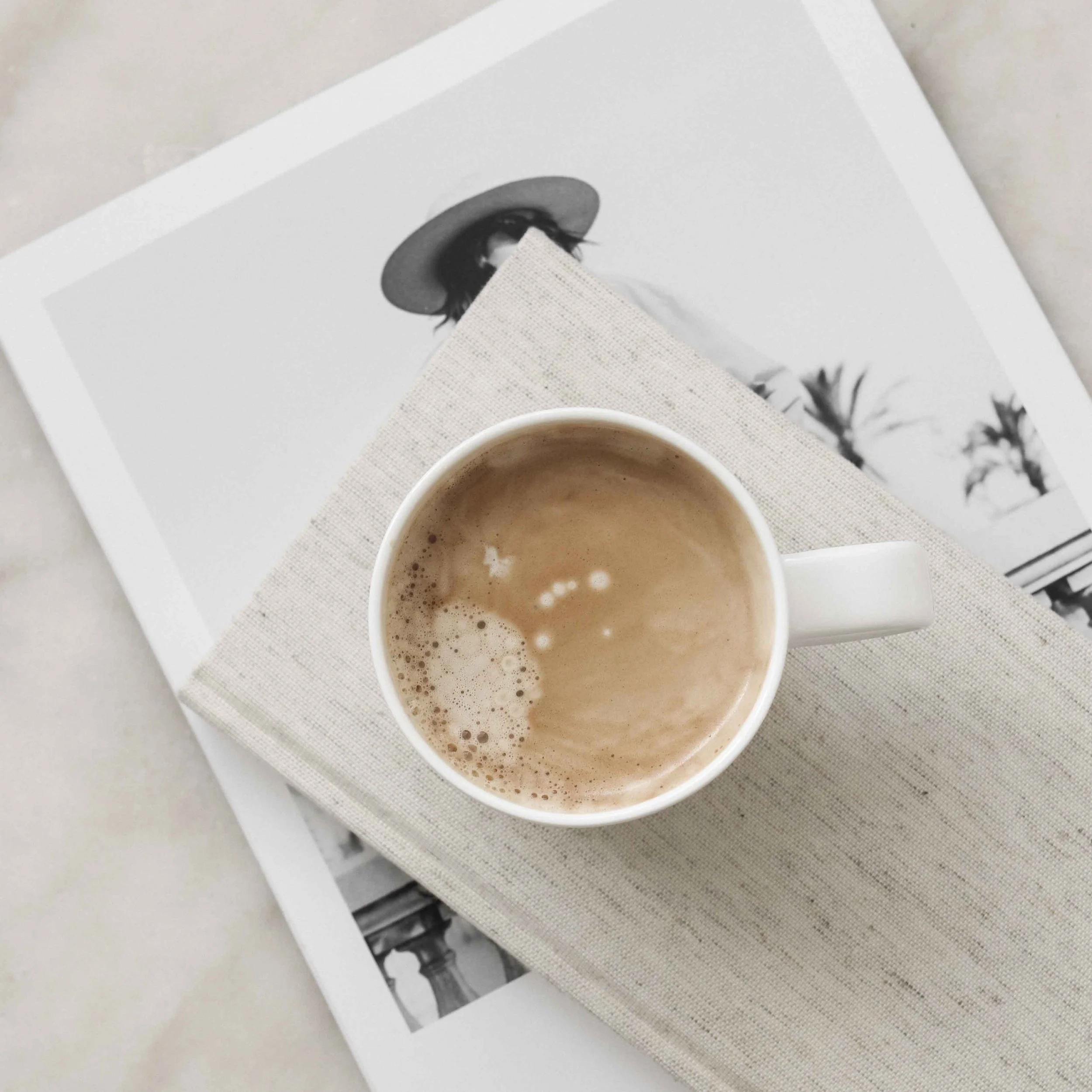 White mug of coffee resting on a linen notebook over a black and white magazine