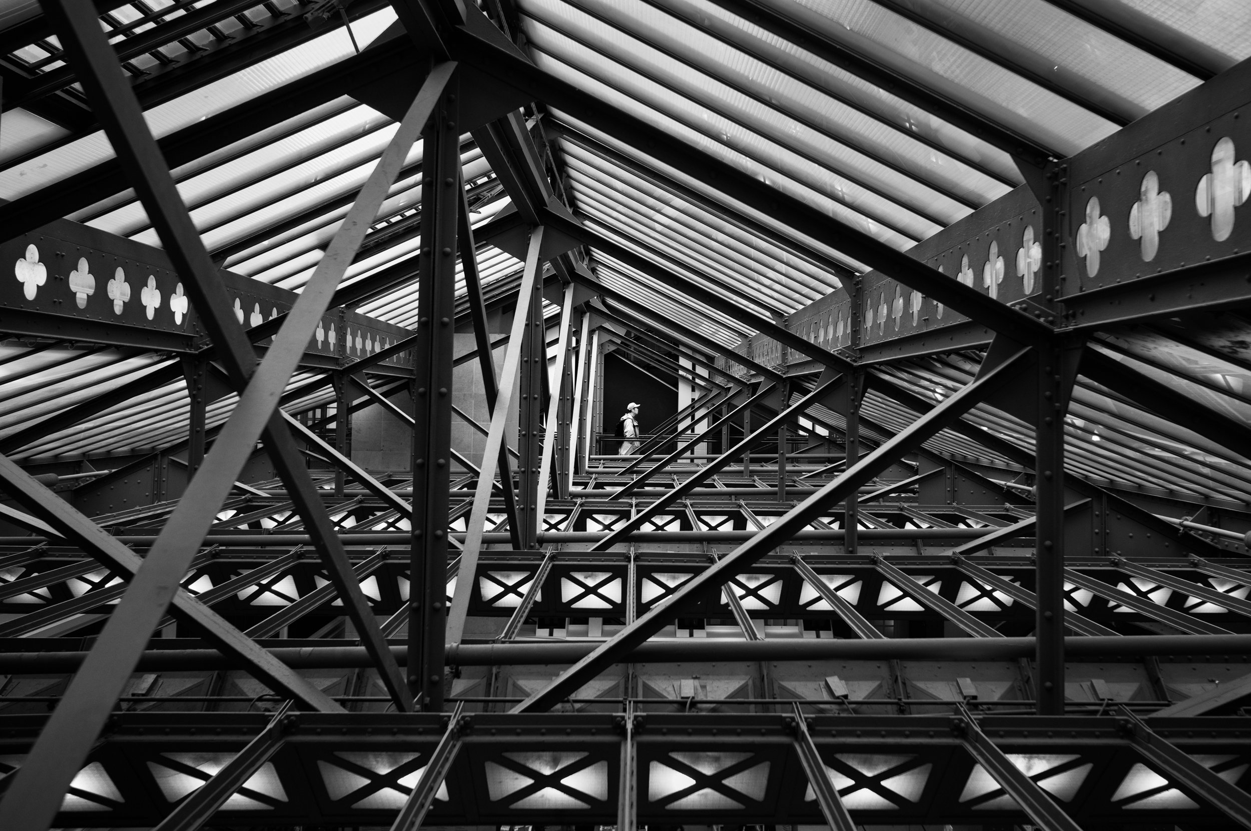 Inside the Eiffel Tower with a person walking on a narrow walkway among metal beams and structural elements, view looking upwards in black and white.