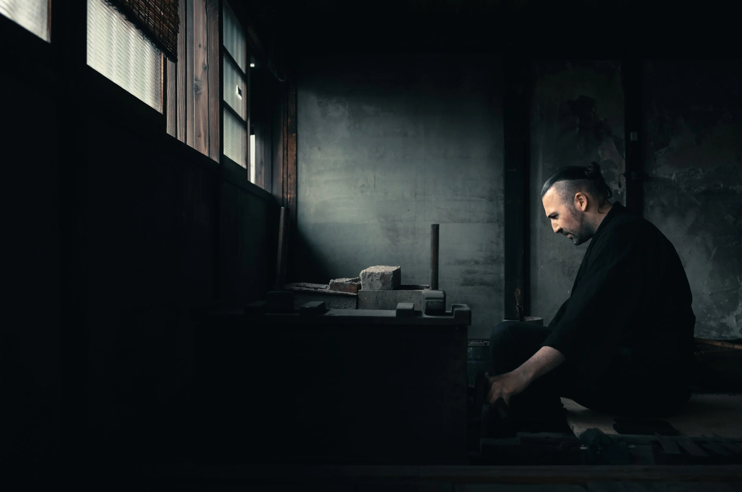 A man dressed in black is sitting on the floor in a dimly lit room with wooden and concrete walls, working on a traditional Japanese tea ceremony clay stove with brick and metal parts.