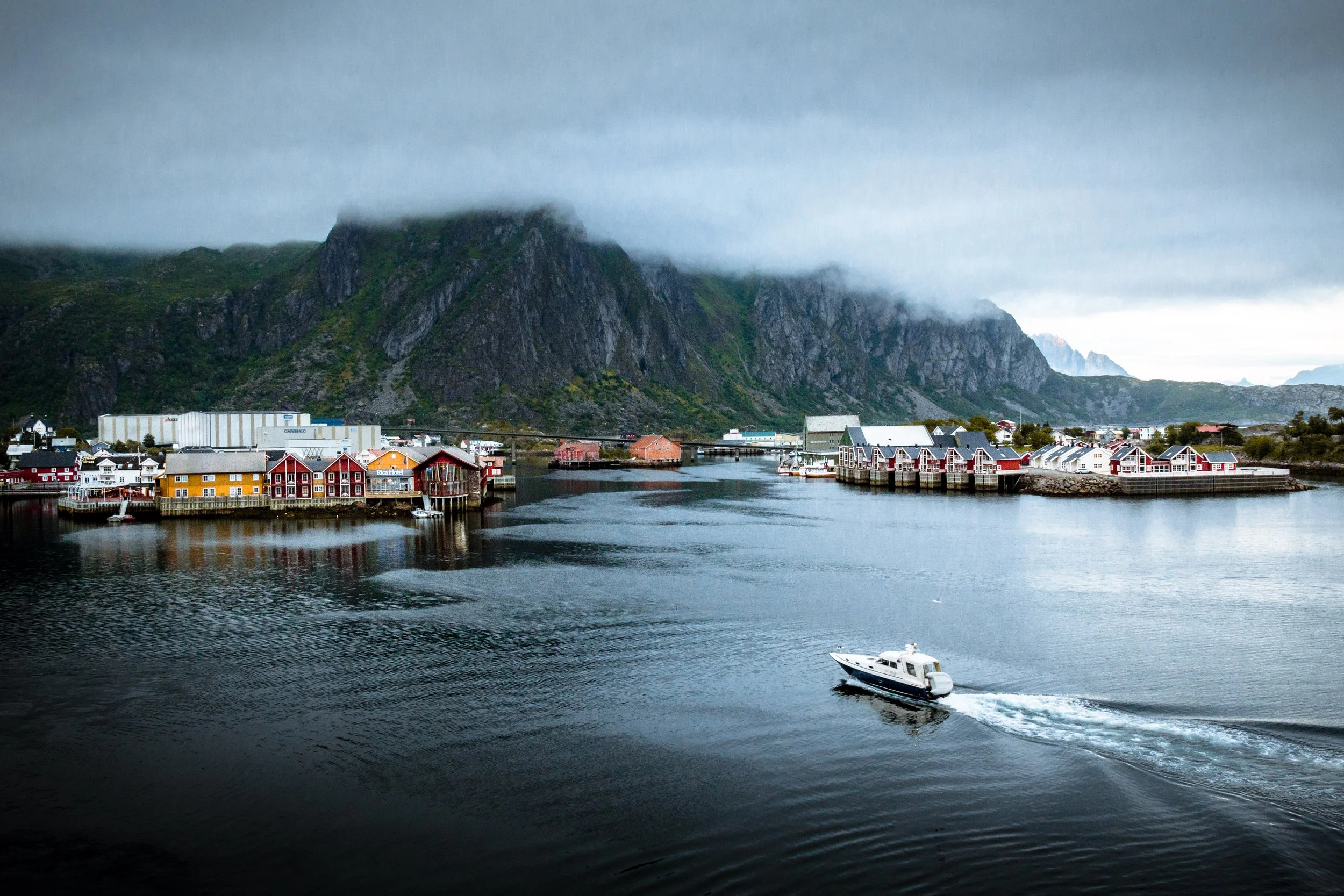 A small boat sailing on calm water with colorful houses on the waterfront and large mountains with clouds in the background.
