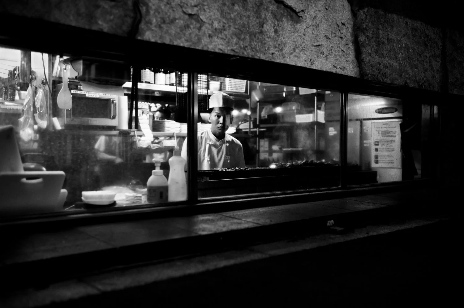 Black and white photo of a chef looking through a restaurant window, with kitchen supplies and equipment inside.