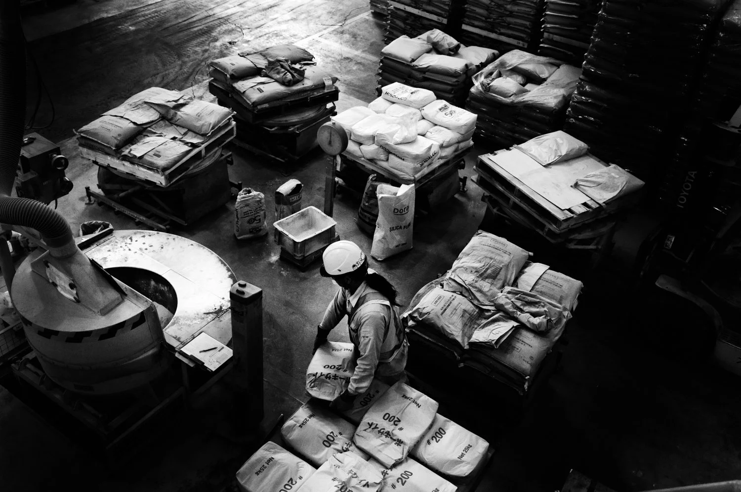 A worker in a safety helmet and vest handling bags of industrial materials in a warehouse with stacks of bags and pallets around.