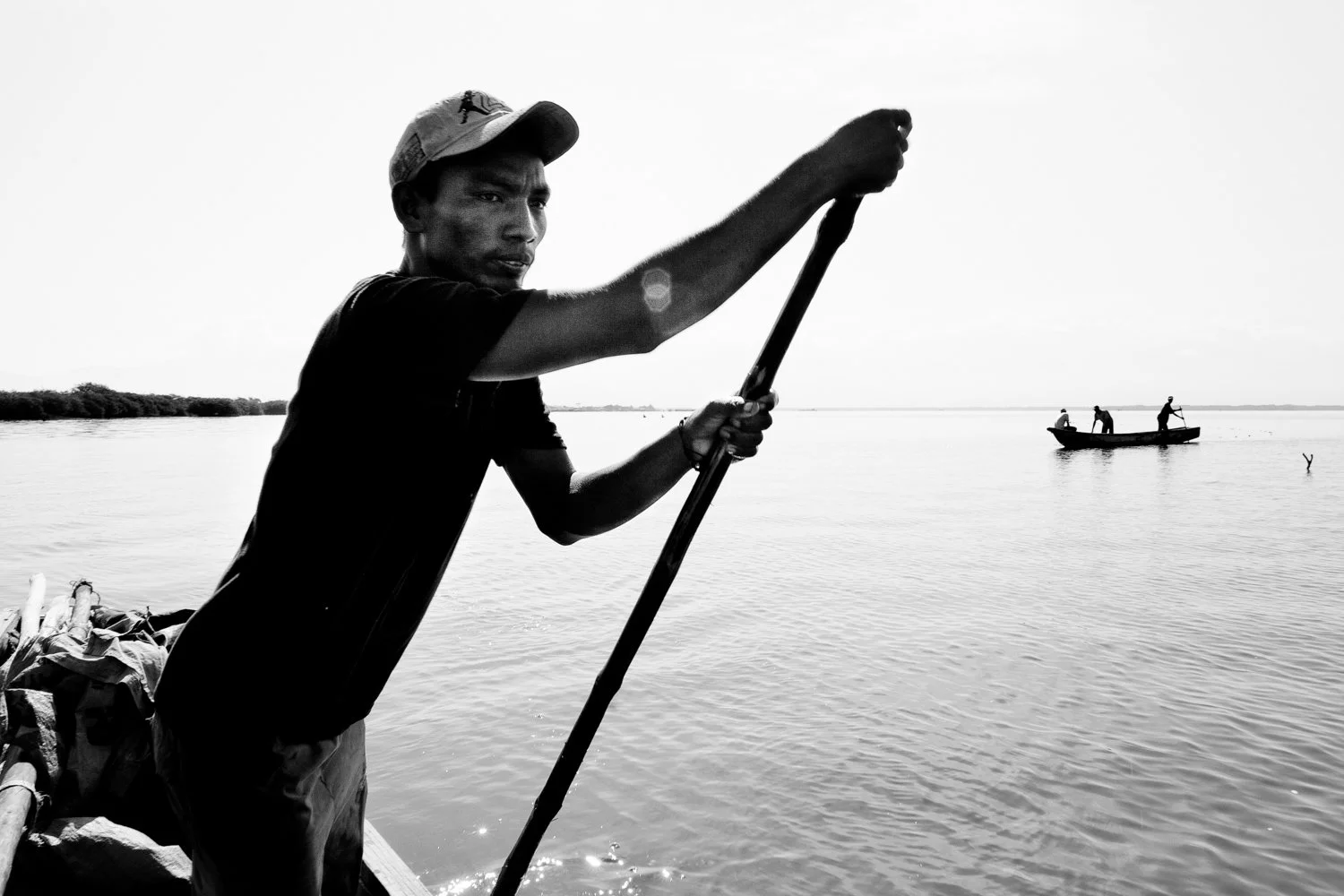 A young man standing on a boat holding a pole, with a body of water and a small boat with three people in the background.