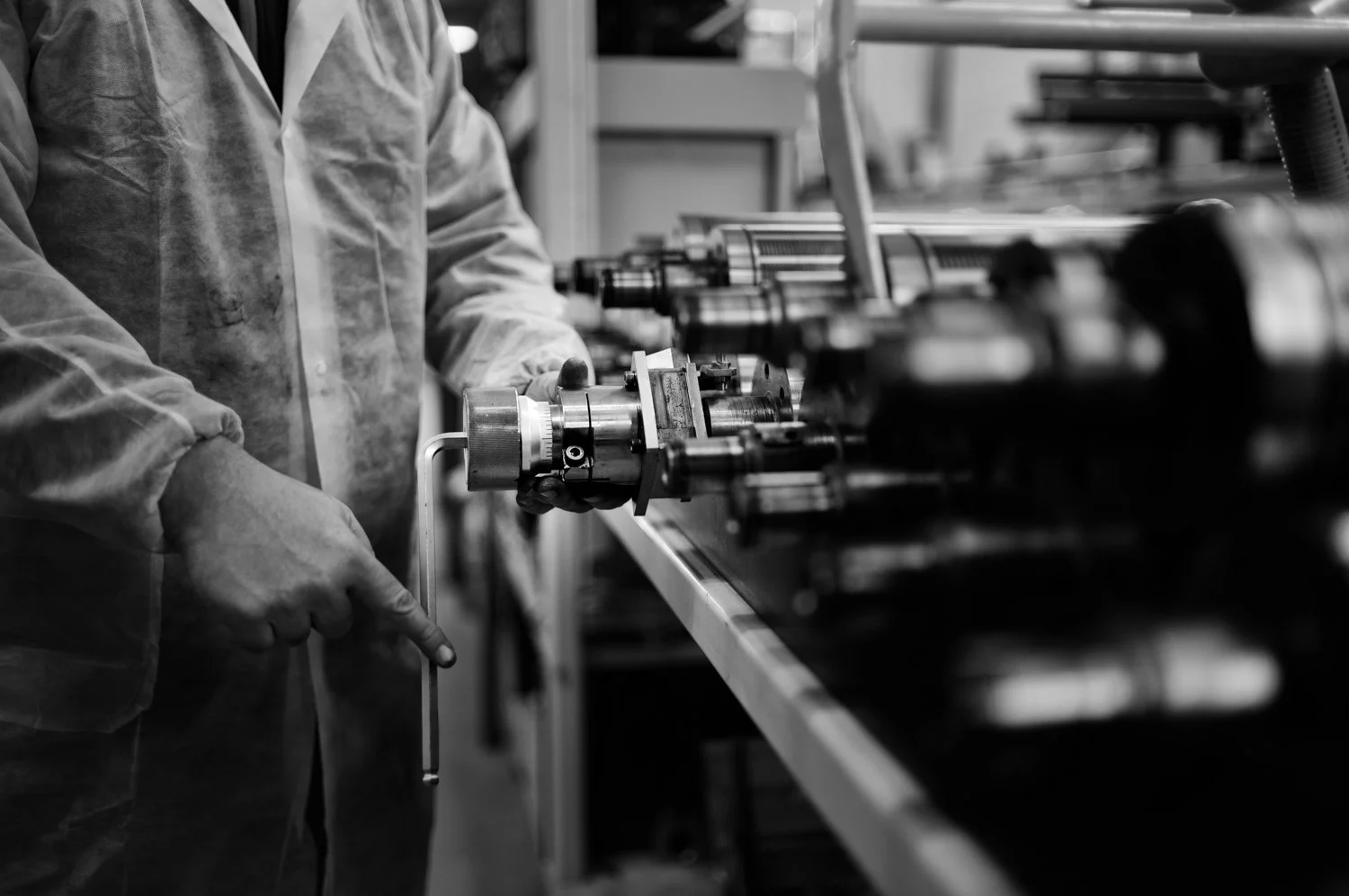 A worker in a factory wearing gloves and a lab coat handles machinery on a workbench, with several mechanical components arranged in a row.