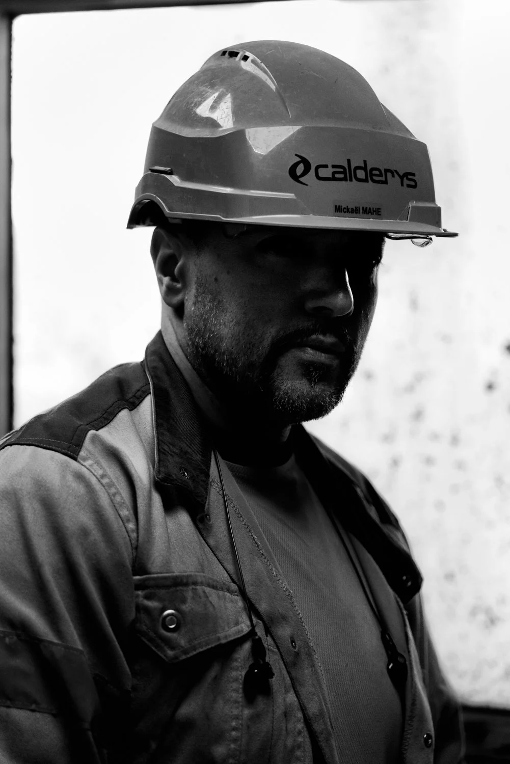 A man wearing a hard hat with the Calders logo and a jacket, standing indoors, in black and white.