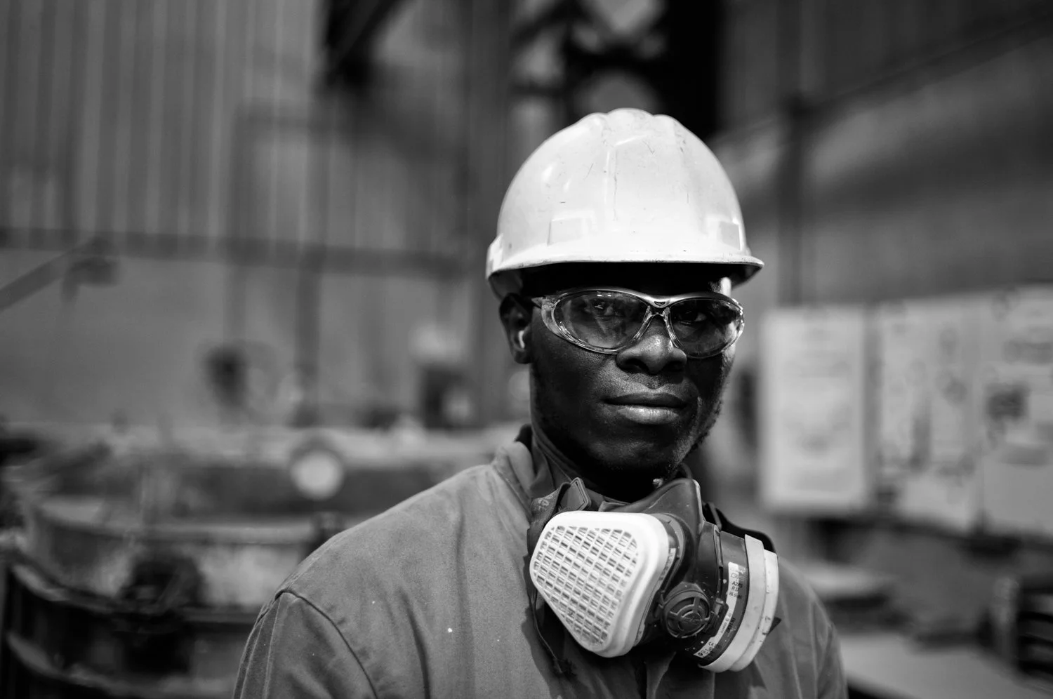 A Black male worker wearing a safety helmet, glasses, and a protective mask around his neck in an industrial setting.