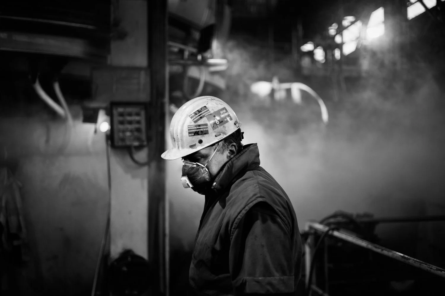 A worker wearing a helmet and face mask, standing in a dimly lit environment with smoke or fog in the background.
