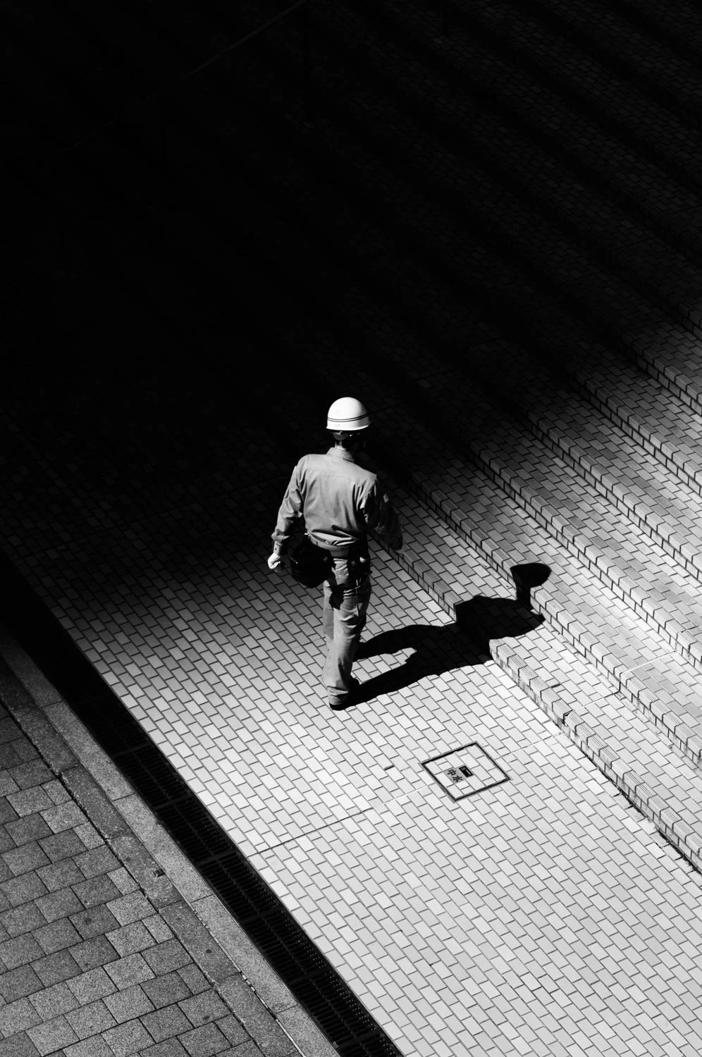 A man wearing a helmet walking on a sidewalk with his shadow cast on the steps, in black and white.