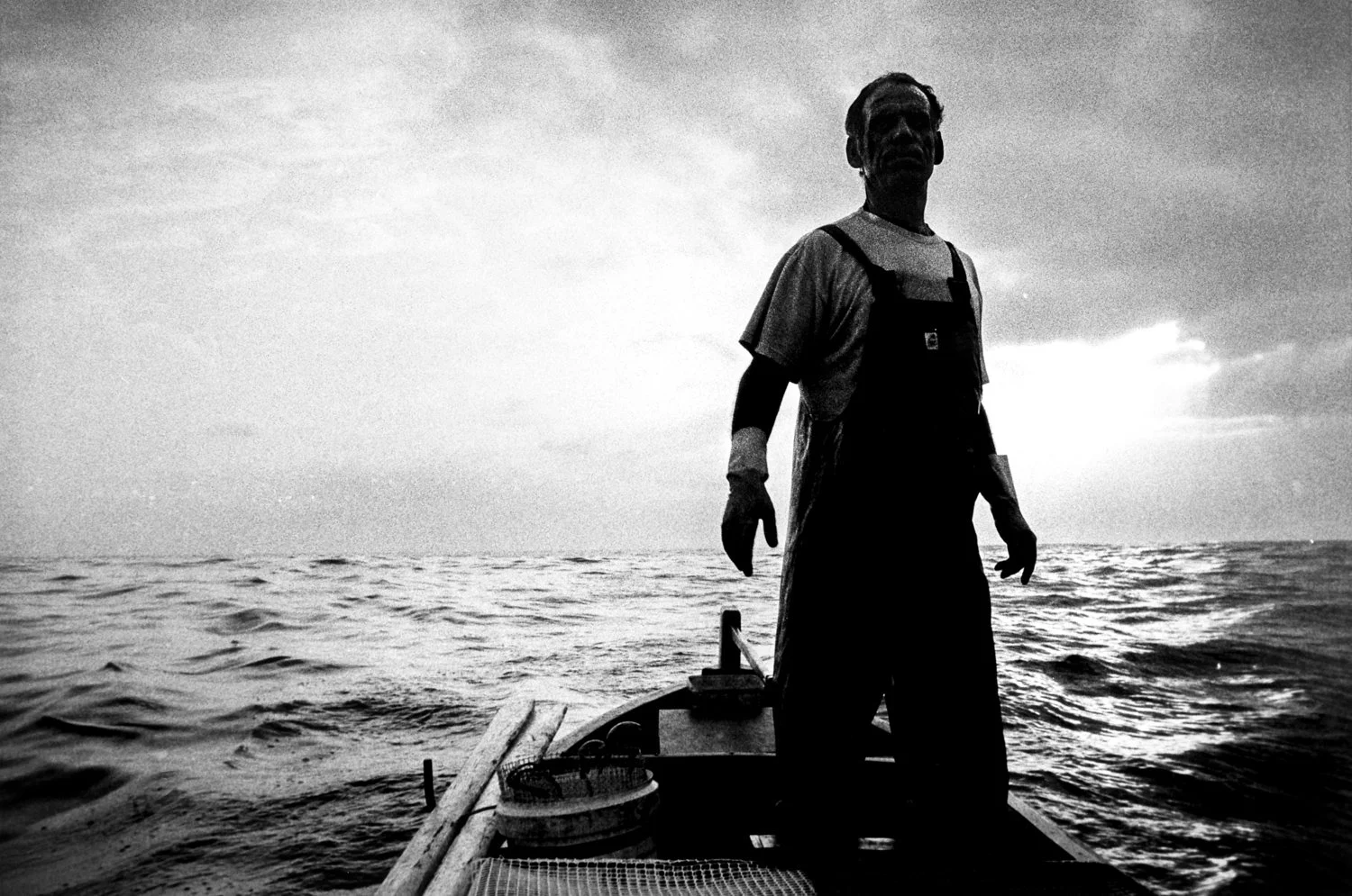 Silhouette of a man standing at the bow of a boat on the water during sunset, with a cloudy sky in the background.