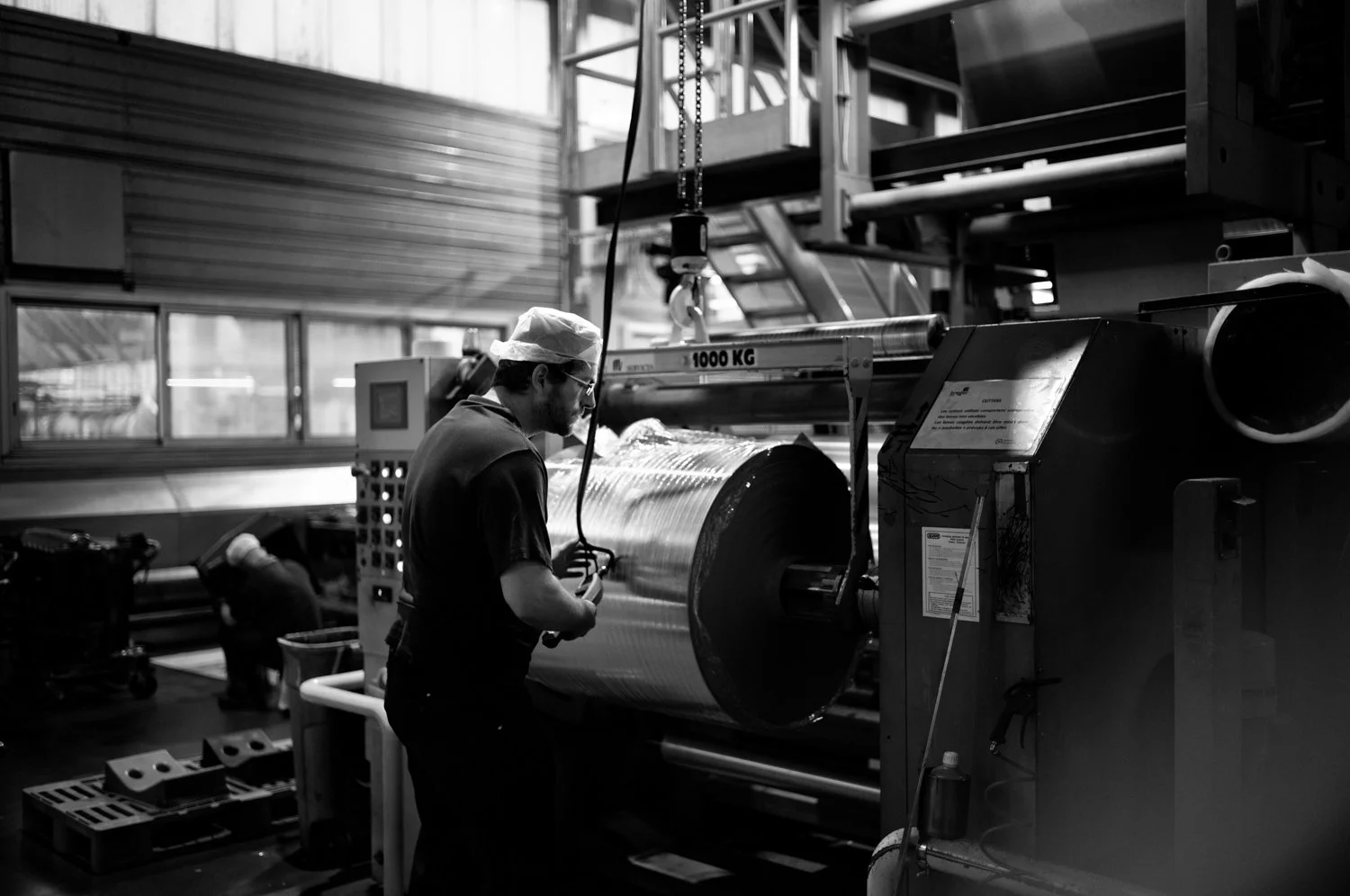 A worker in a factory operates industrial machinery, with a large roll of material and various controls and equipment around.