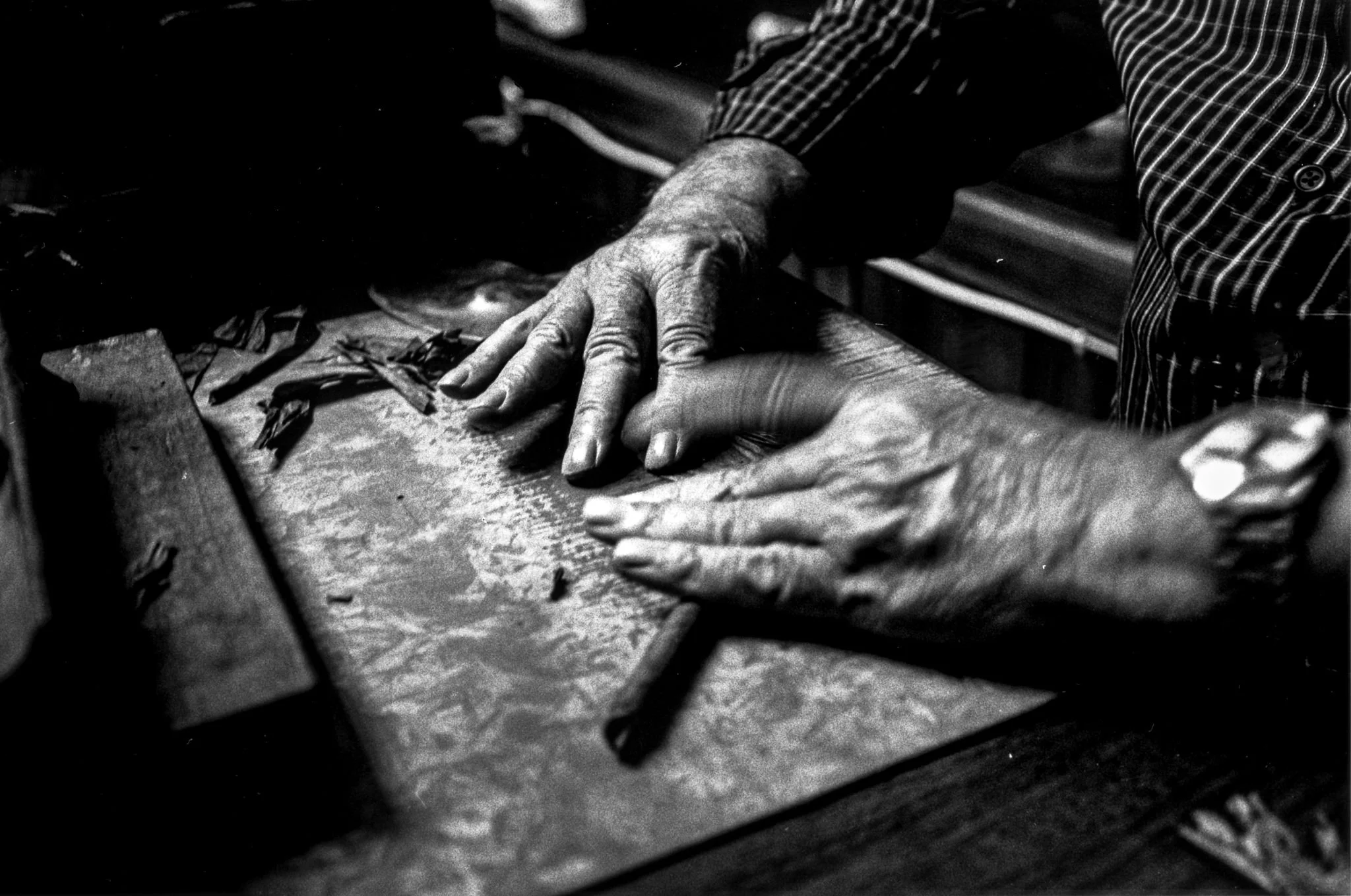 Two elderly hands with prominent veins and wrinkles, one resting on a wooden surface and the other reaching toward it, in a black and white photo.