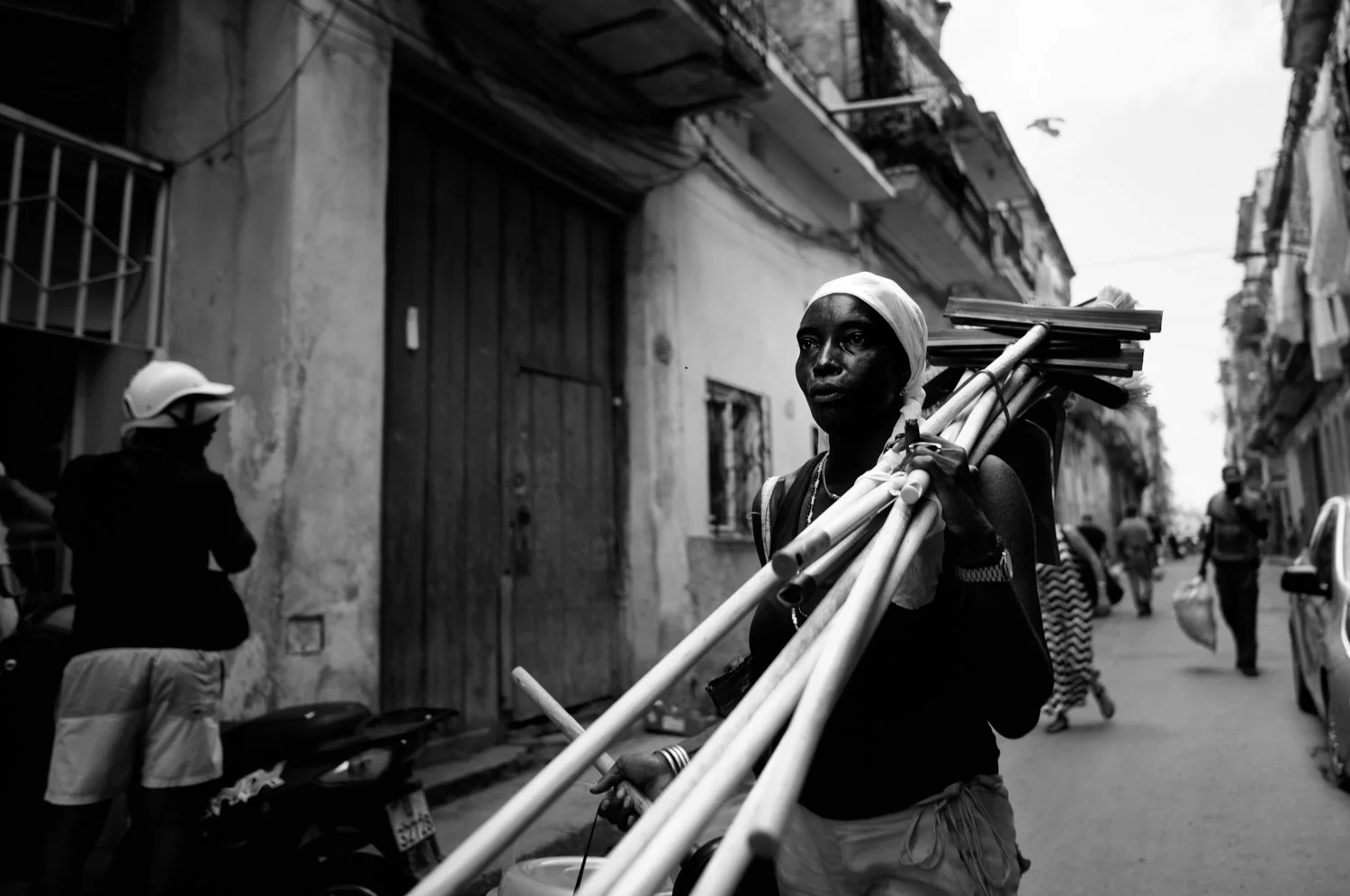 A woman walking down a street carrying a bundle of long cylindrical objects, with other people and buildings visible in the background.