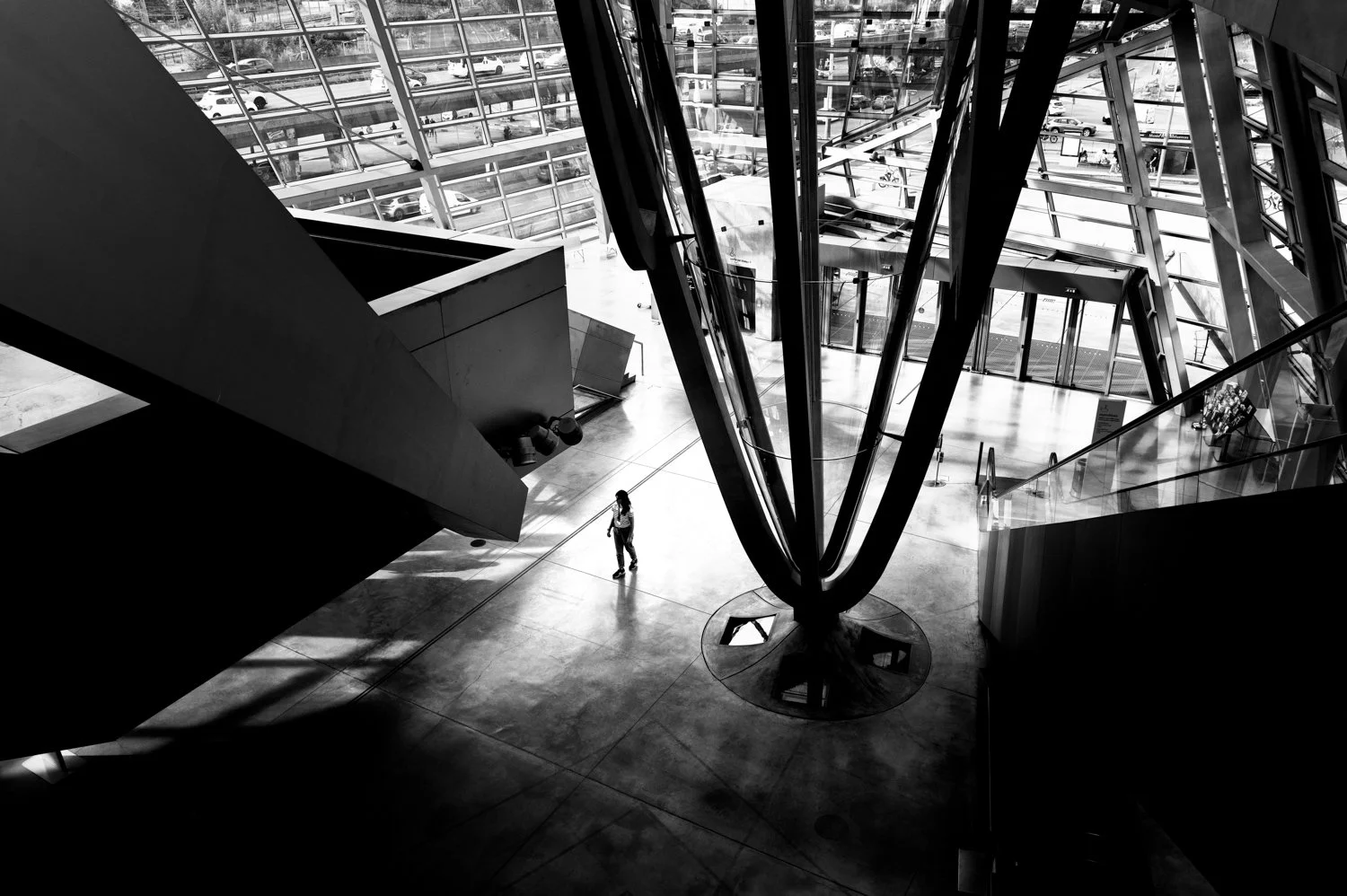Black and white photo of the interior of a modern building with large glass windows. There is a person walking on the floor near the center, and a prominent vertical structure with glass and metal elements.