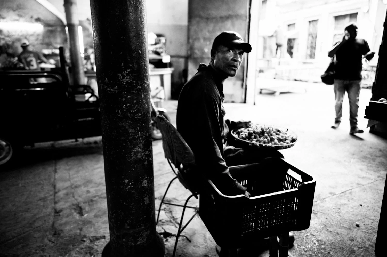 A black and white photo of a man sitting with a cart, holding a basket of fruit or vegetables, in an urban outdoor setting with a pole in the foreground and people walking in the background.