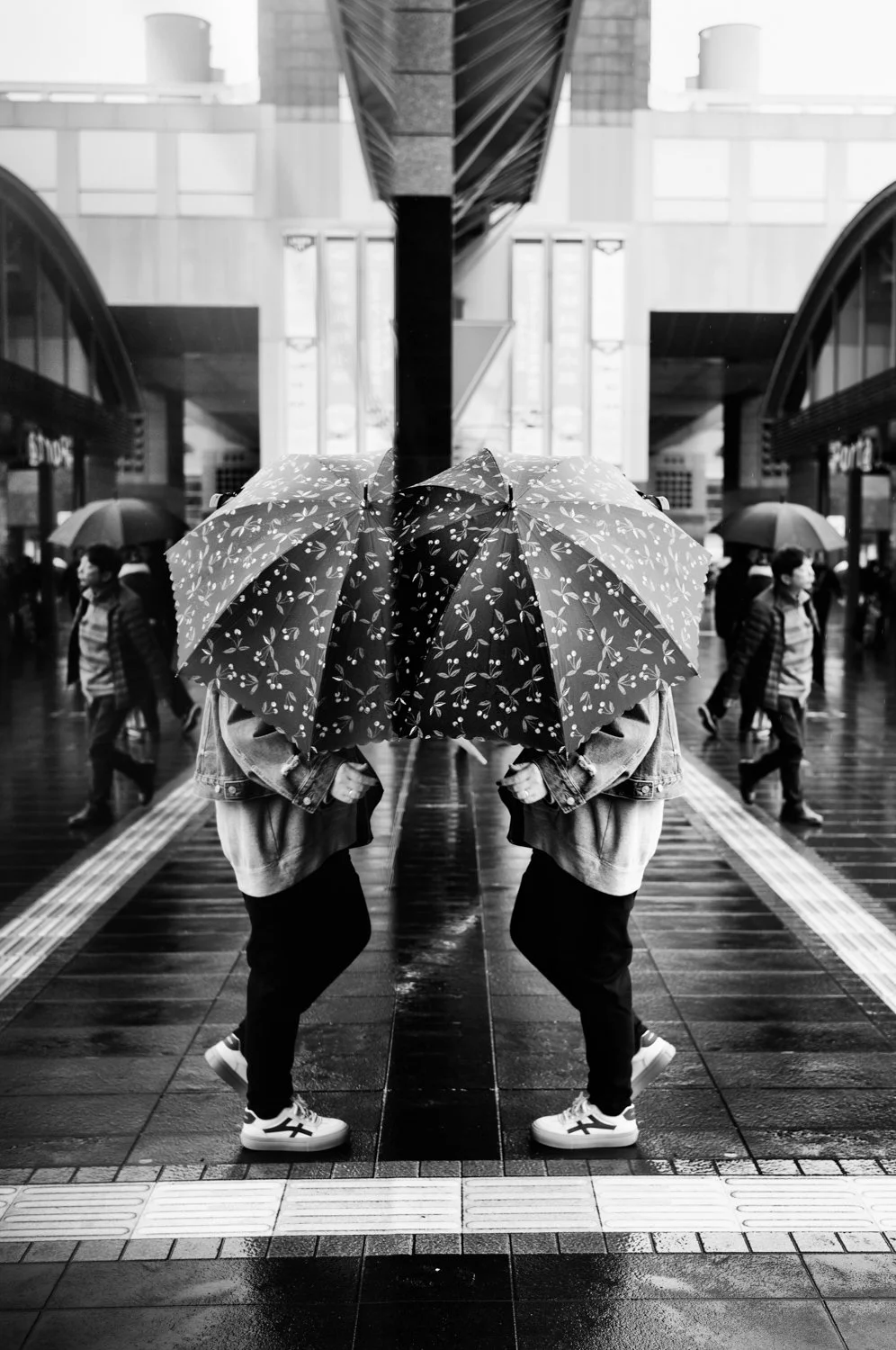 Black and white photo of two people standing under umbrellas on a rainy day in an urban area, with reflections on the wet pavement and passersby in the background.