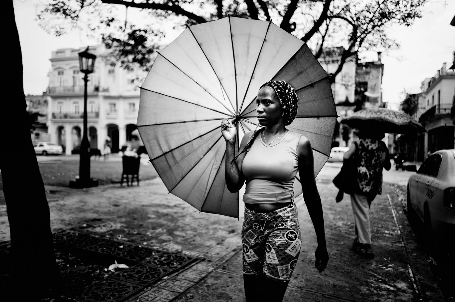 A woman walking on a city street holding an umbrella, with other people and buildings in the background, in black and white.