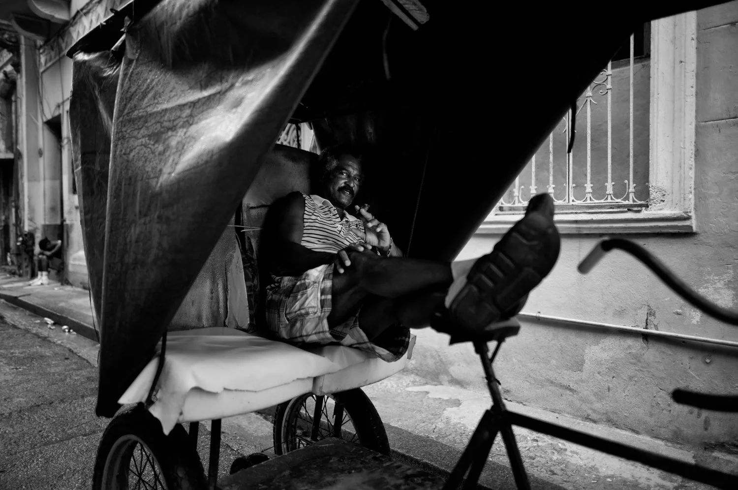 A man sitting in a rickshaw on a city street, holding a cigar, wearing a striped shirt and shorts, with a window and railing seen in the background.
