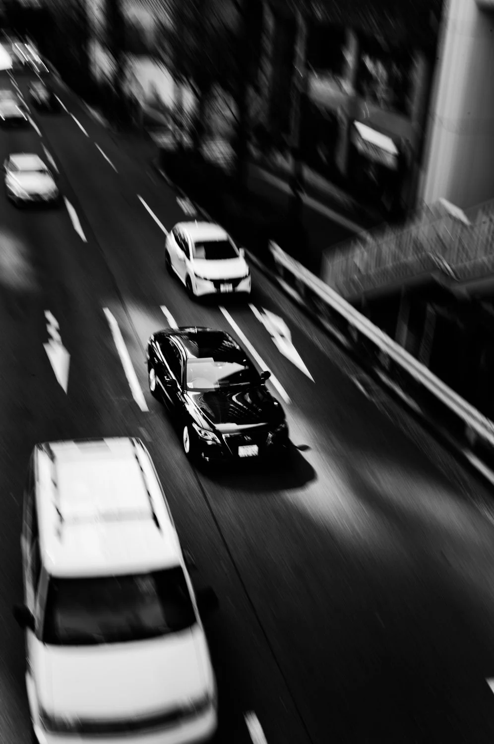 Black car driving on city street with other vehicles parked and moving in black and white urban scene.
