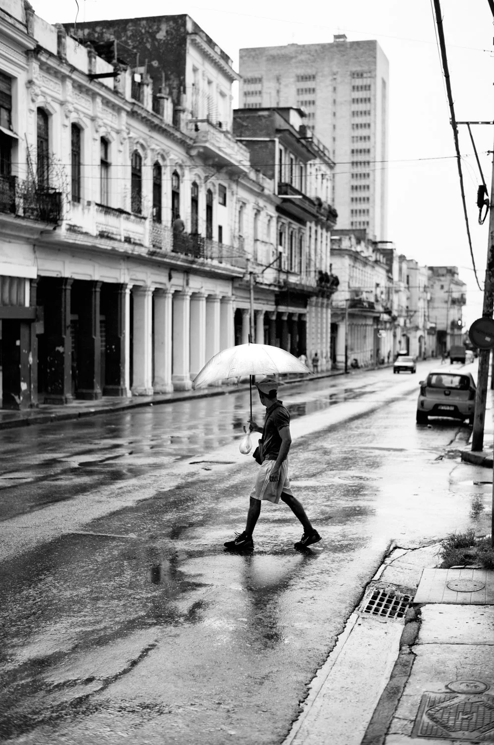 A man crossing a wet city street with an umbrella during rain, with historic buildings and cars in the background.