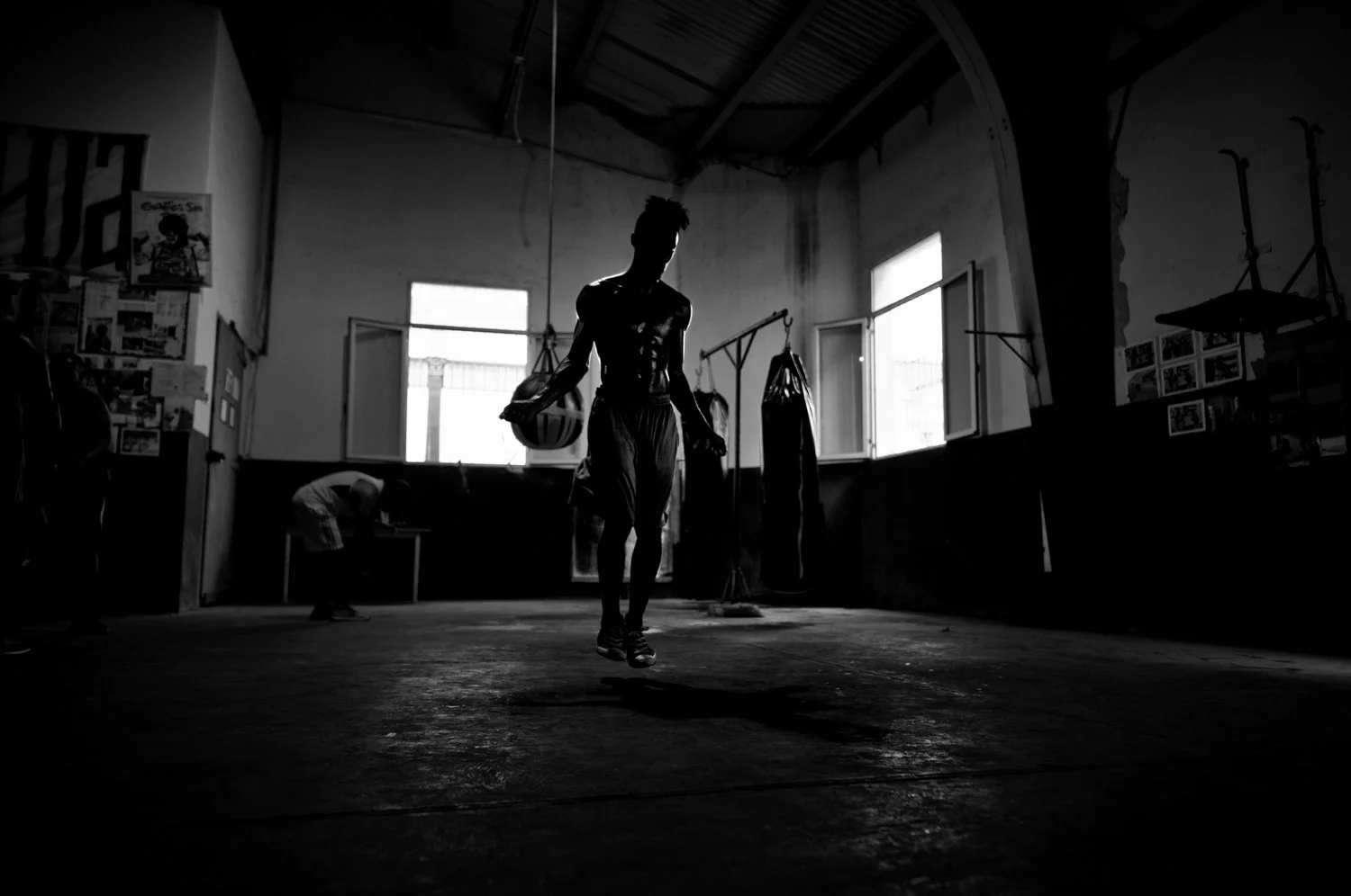 A person jumped rope in a boxing gym with boxing equipment hanging in the background, silhouette of a person with bright light from windows, black and white photo.