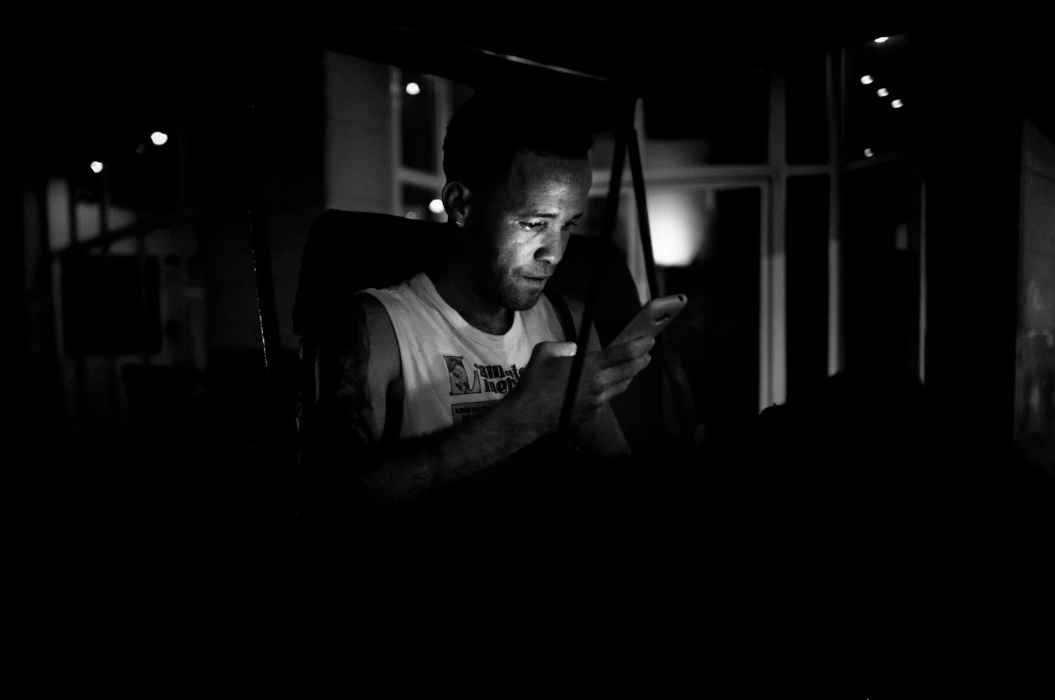 A man is sitting in a dark room, illuminated by the light from his smartphone, with a focused expression.