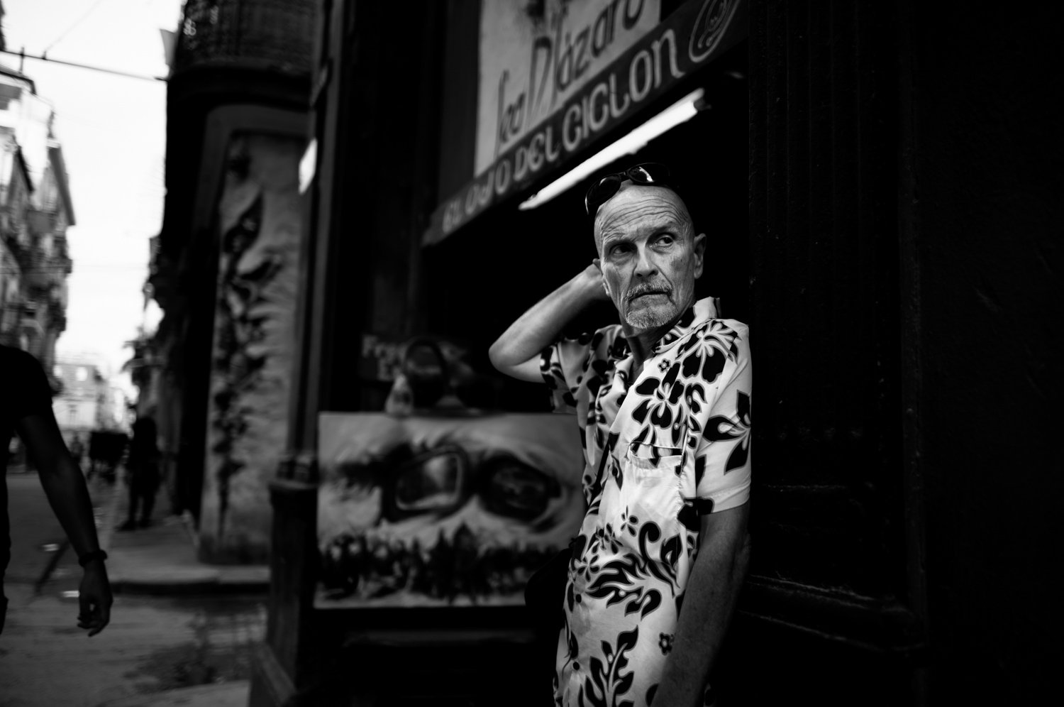 An elderly man wearing sunglasses and a patterned shirt leaning against a building on a busy street, with people walking in the background in black and white.