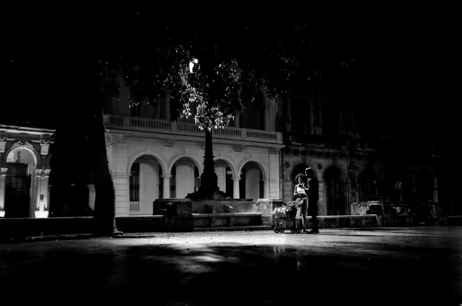 A black-and-white photo of a couple with a baby stroller in a park at night, illuminated by a streetlamp, with a historic building in the background.