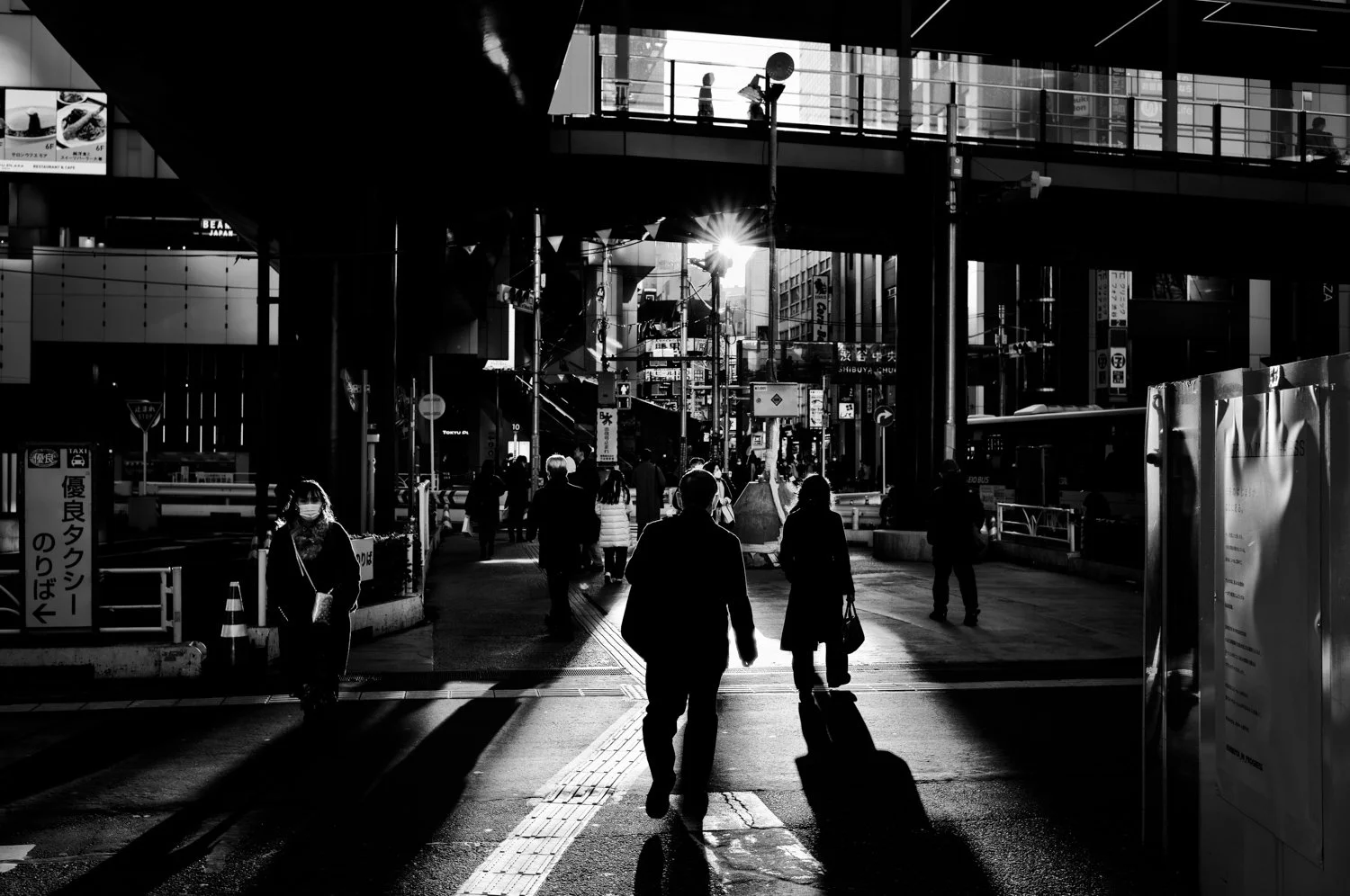 Black and white photo of a city street intersection with people walking, some wearing masks, sunlight shining through buildings, overhead walkways, and various signs and advertisements.